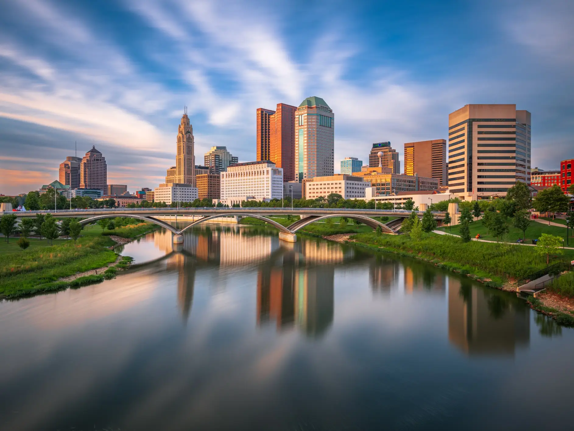 Downtown Columbus skyline and arched bridge reflect in the Scioto River under a streaked evening sky.