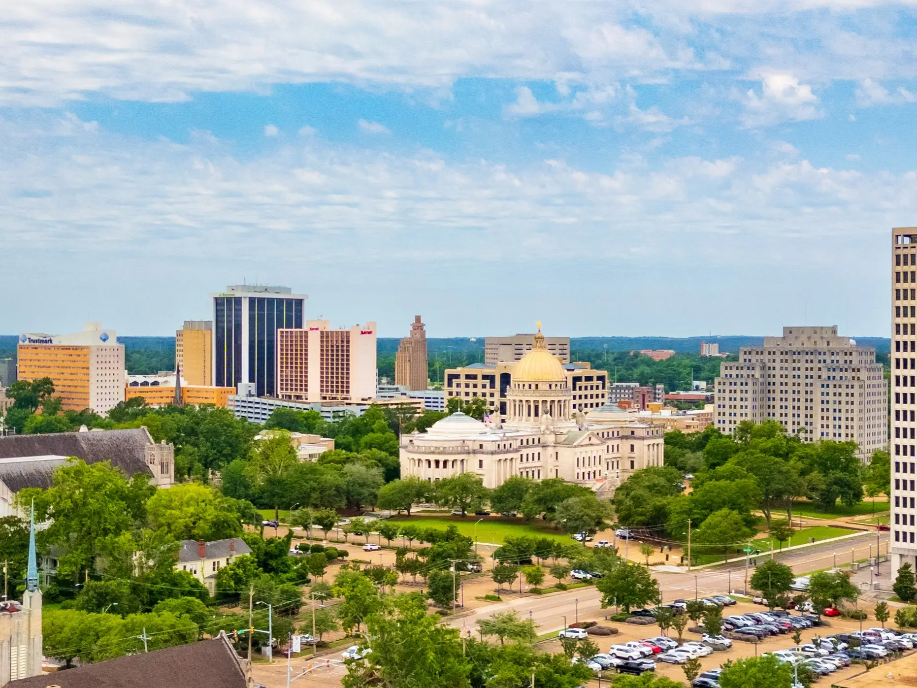 Mississippi State Capitol with a gold dome stands among trees and downtown Jackson high-rises under a cloudy sky.