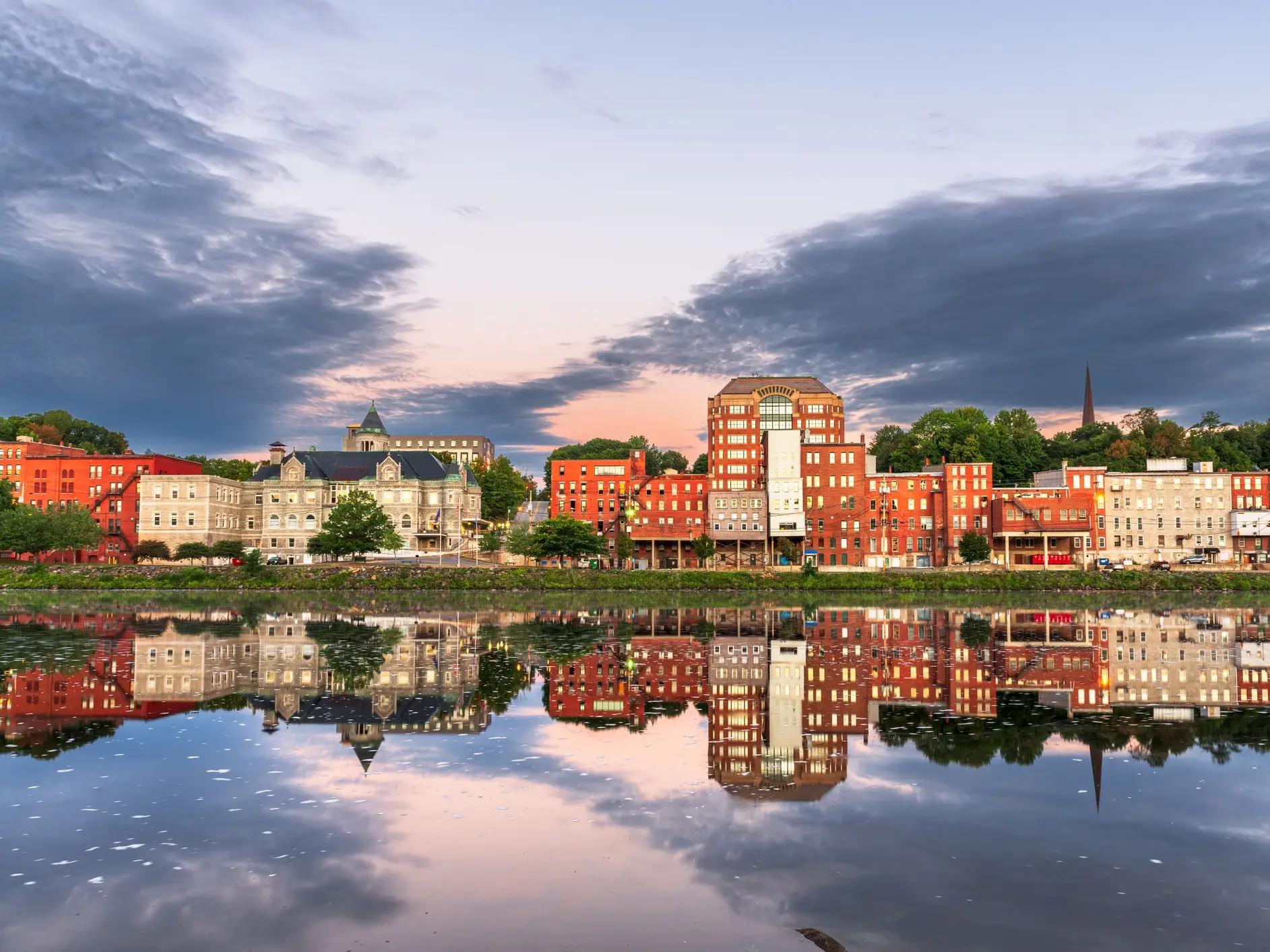 Waterfront brick buildings and a church spire reflect in a calm river beneath a cloudy dusk sky.