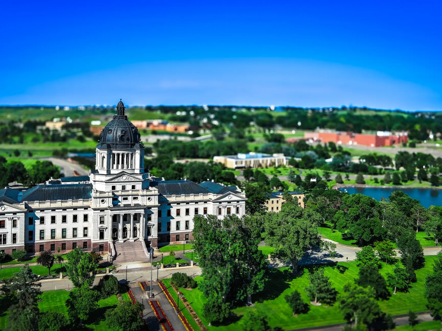 Aerial view of the South Dakota State Capitol with a domed roof, surrounding lawns, trees, and nearby water.