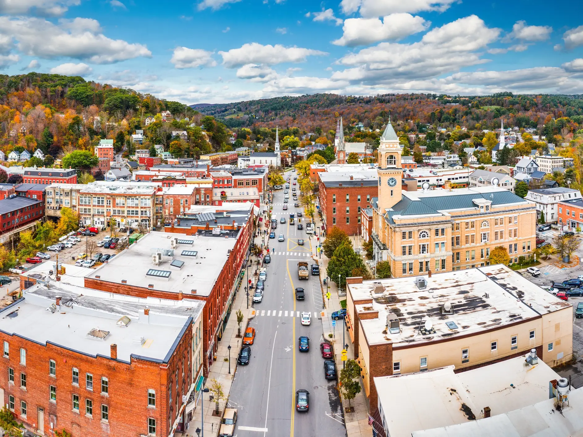 Aerial view of a downtown street lined with brick buildings, a clock tower, cars, and autumn hills.