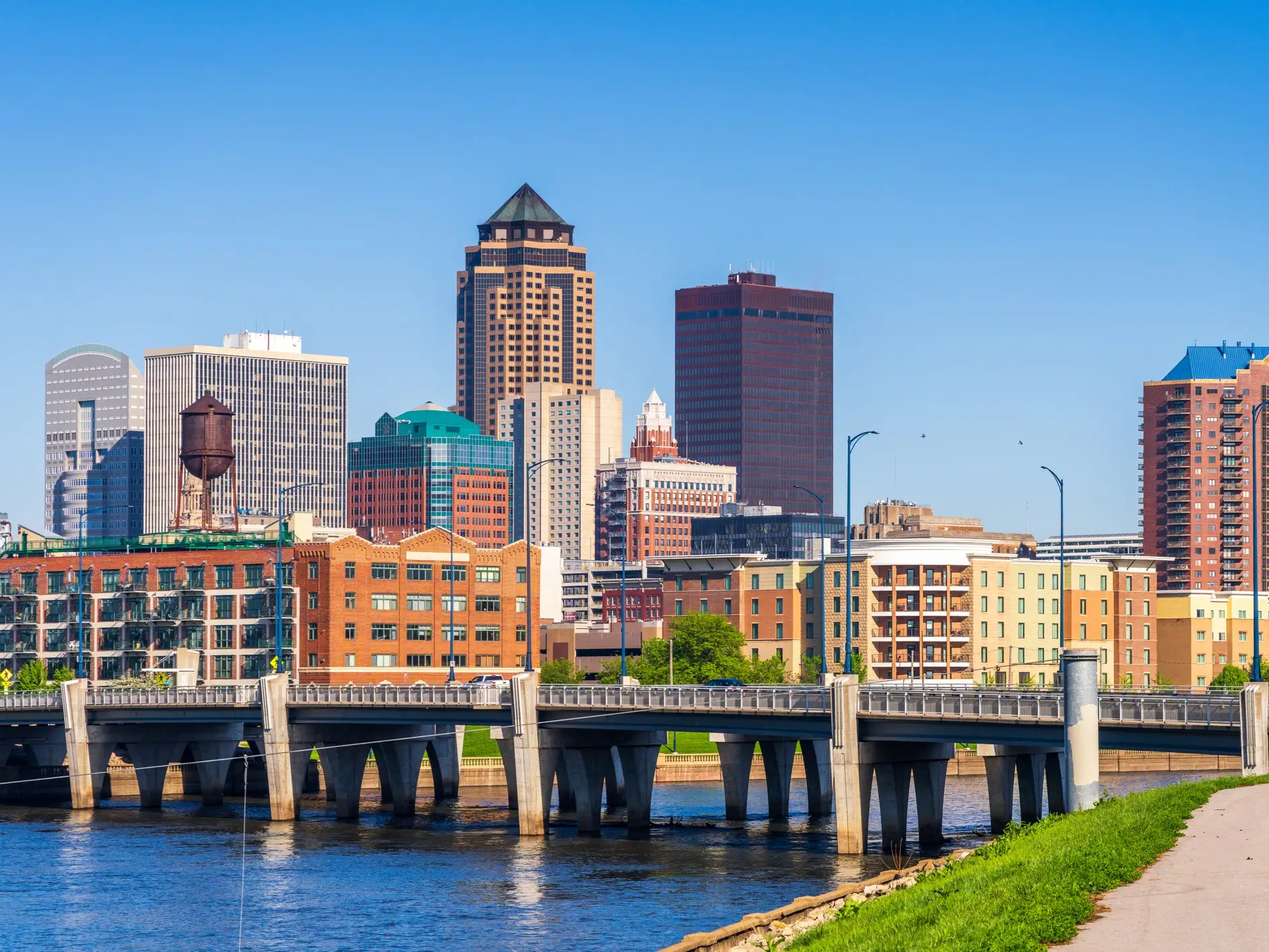 Downtown Des Moines skyline rises behind a bridge over the river on a clear sunny day.