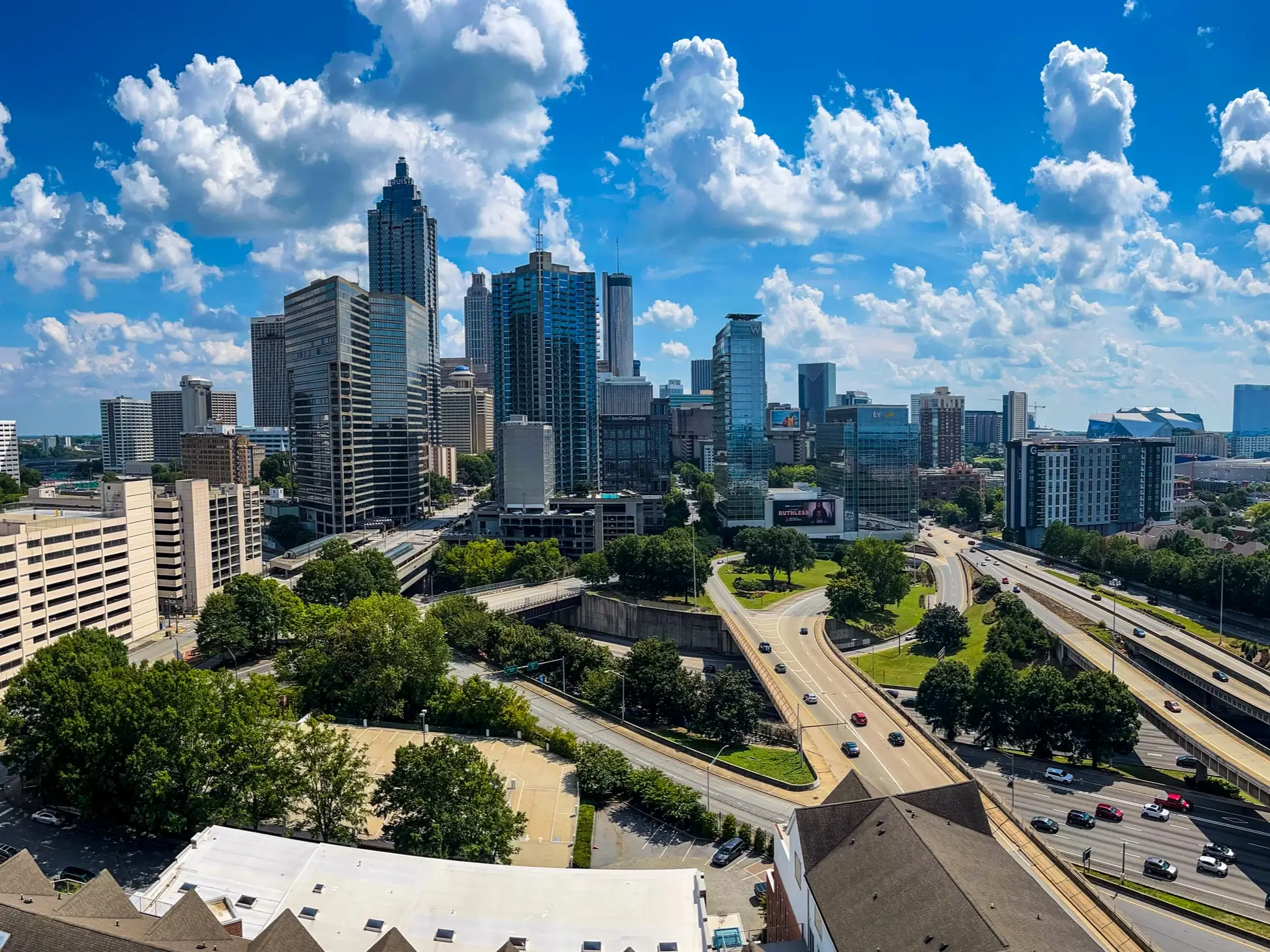 Downtown Atlanta skyline rises above highways and trees under a bright blue sky with large cumulus clouds.