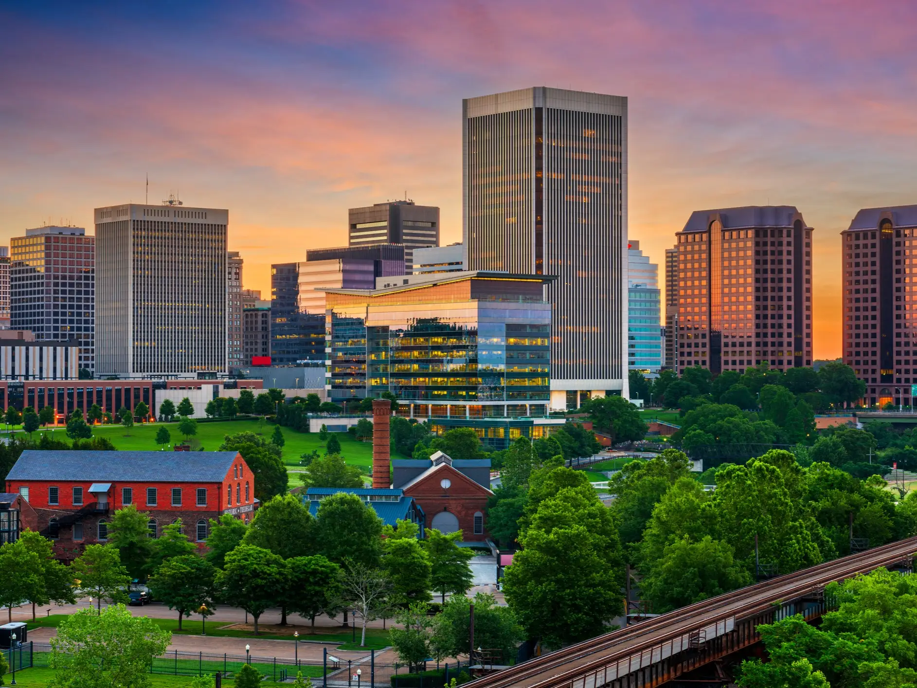 Downtown Richmond skyline rises above green parkland and brick buildings under a colorful sunset sky.
