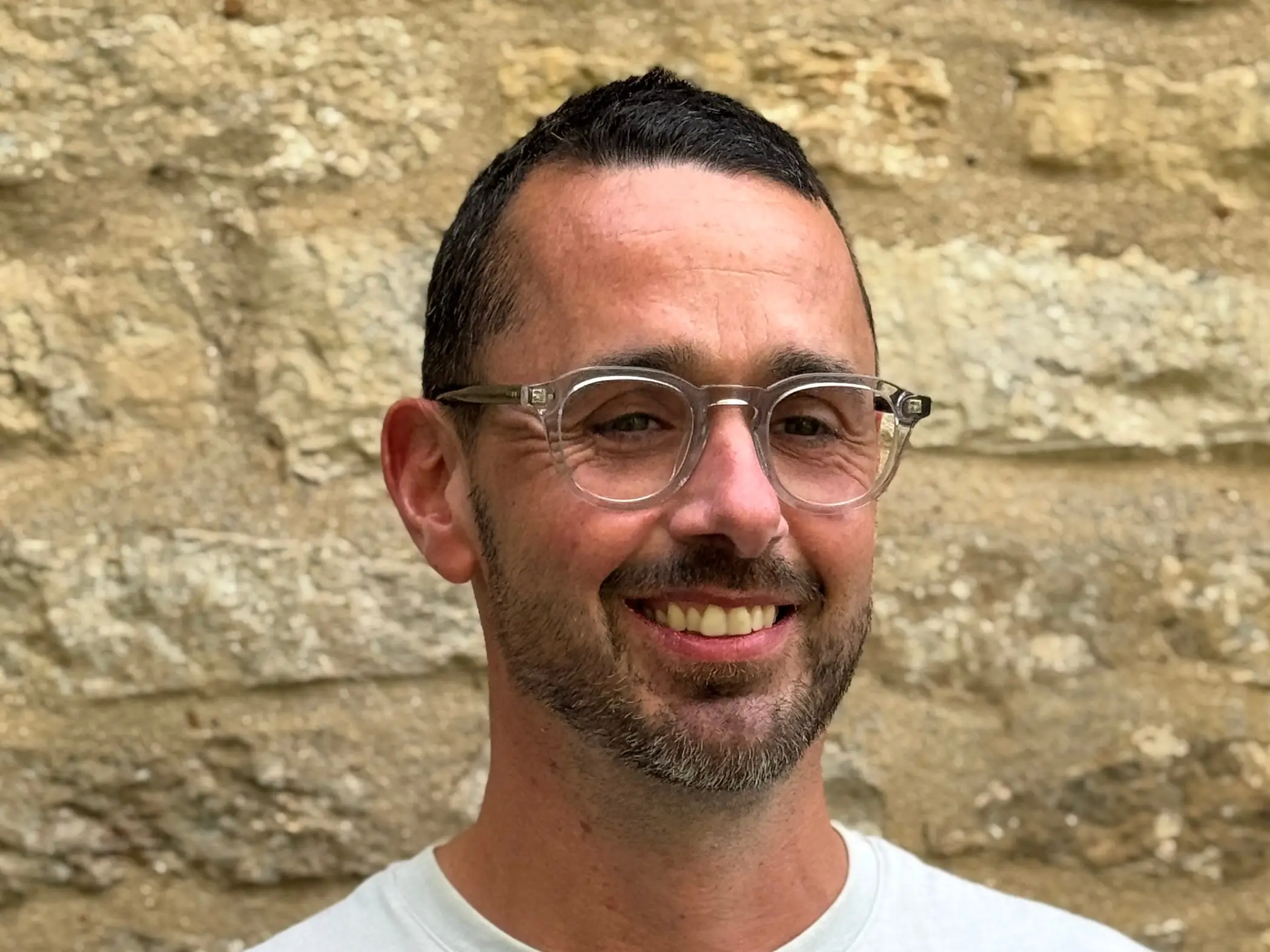 Smiling person with clear glasses and a light shirt stands in front of a stone wall.