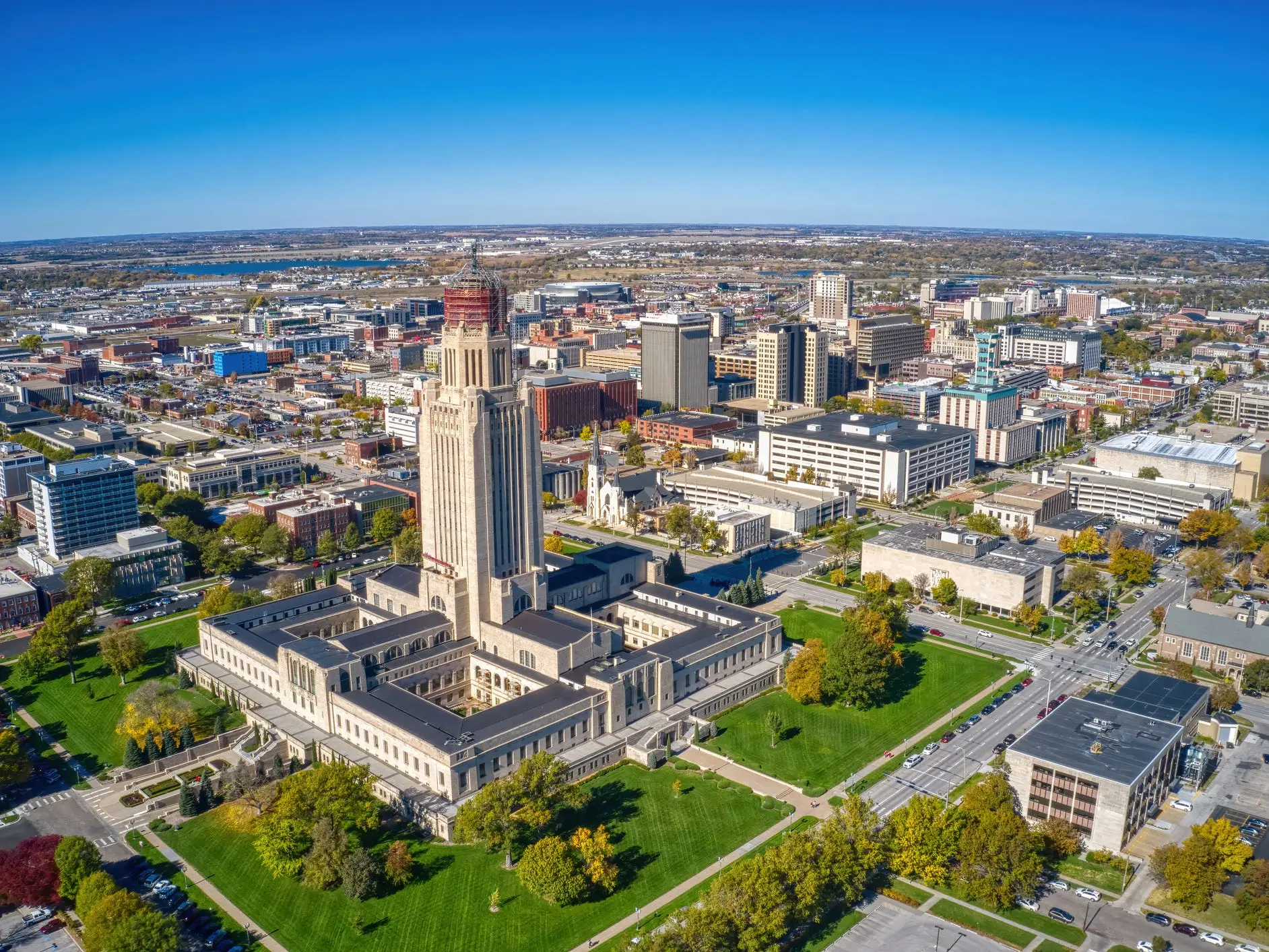 Aerial view of the Nebraska State Capitol surrounded by downtown Lincoln streets, buildings, lawns, and autumn trees.