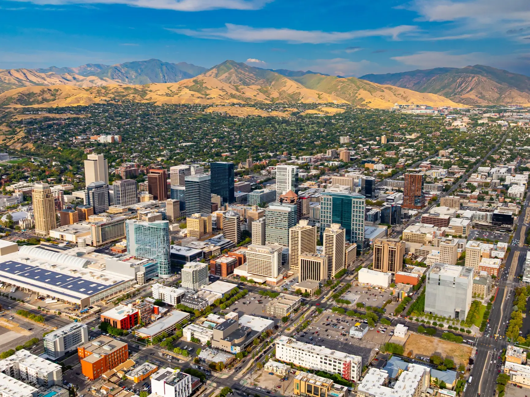 Downtown Salt Lake City skyline spreads toward the Wasatch Mountains under a partly cloudy blue sky.