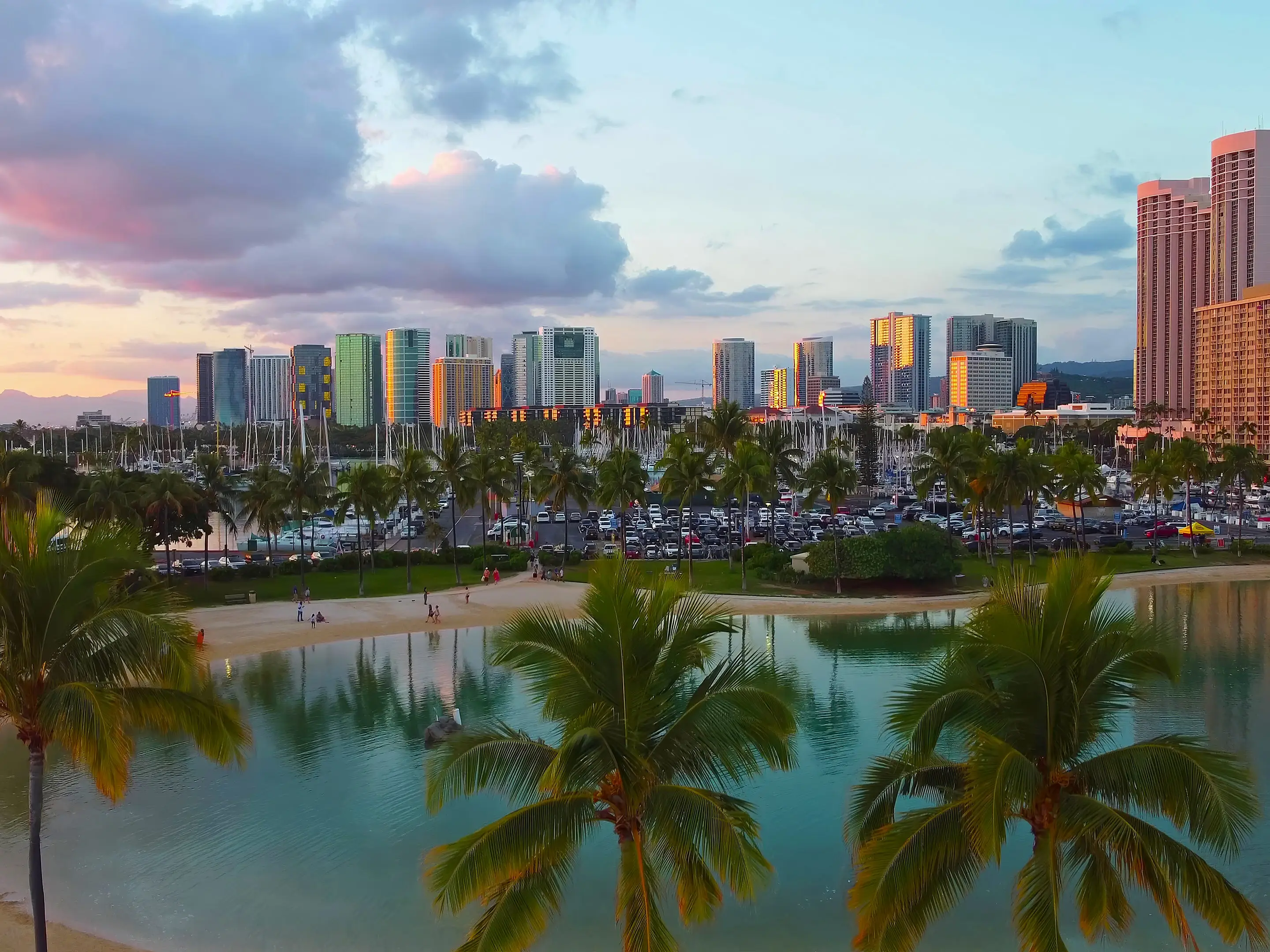 Palm trees frame a waterfront lagoon, marina, and high-rise skyline under colorful sunset clouds in Honolulu.
