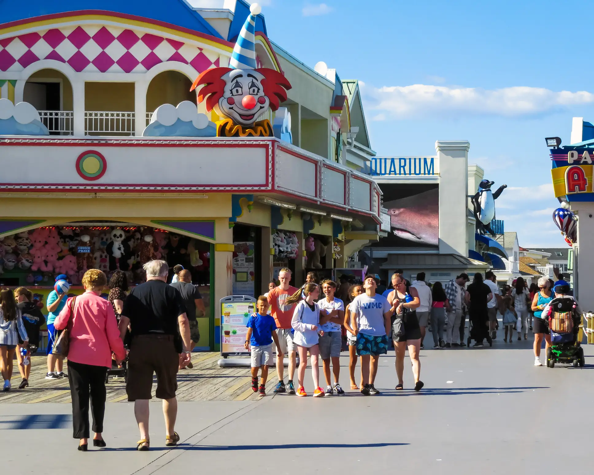 Crowds walk along a sunny amusement boardwalk past a colorful clown-themed arcade and aquarium sign.