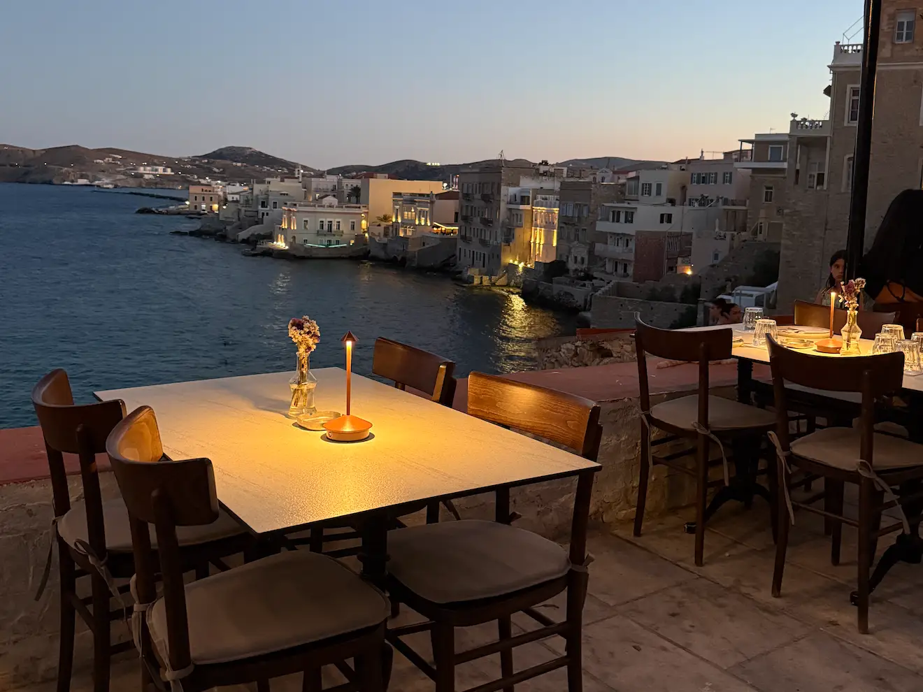Outdoor restaurant terrace with candlelit tables in Syros overlooking the water and harbor at dusk.
