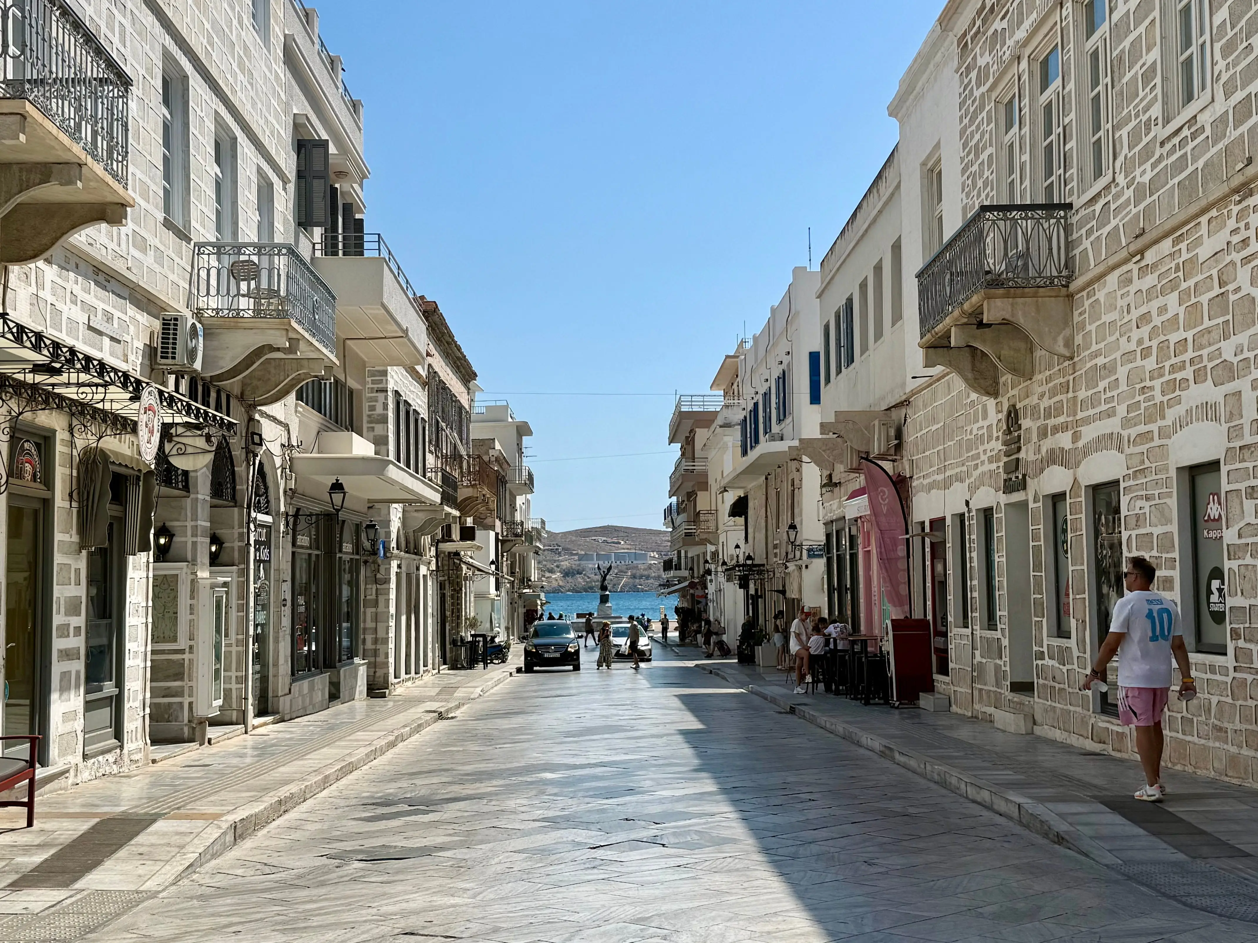 A quiet sunlit stone street lined with balconies and shops leads toward the blue harbor water in Syros.