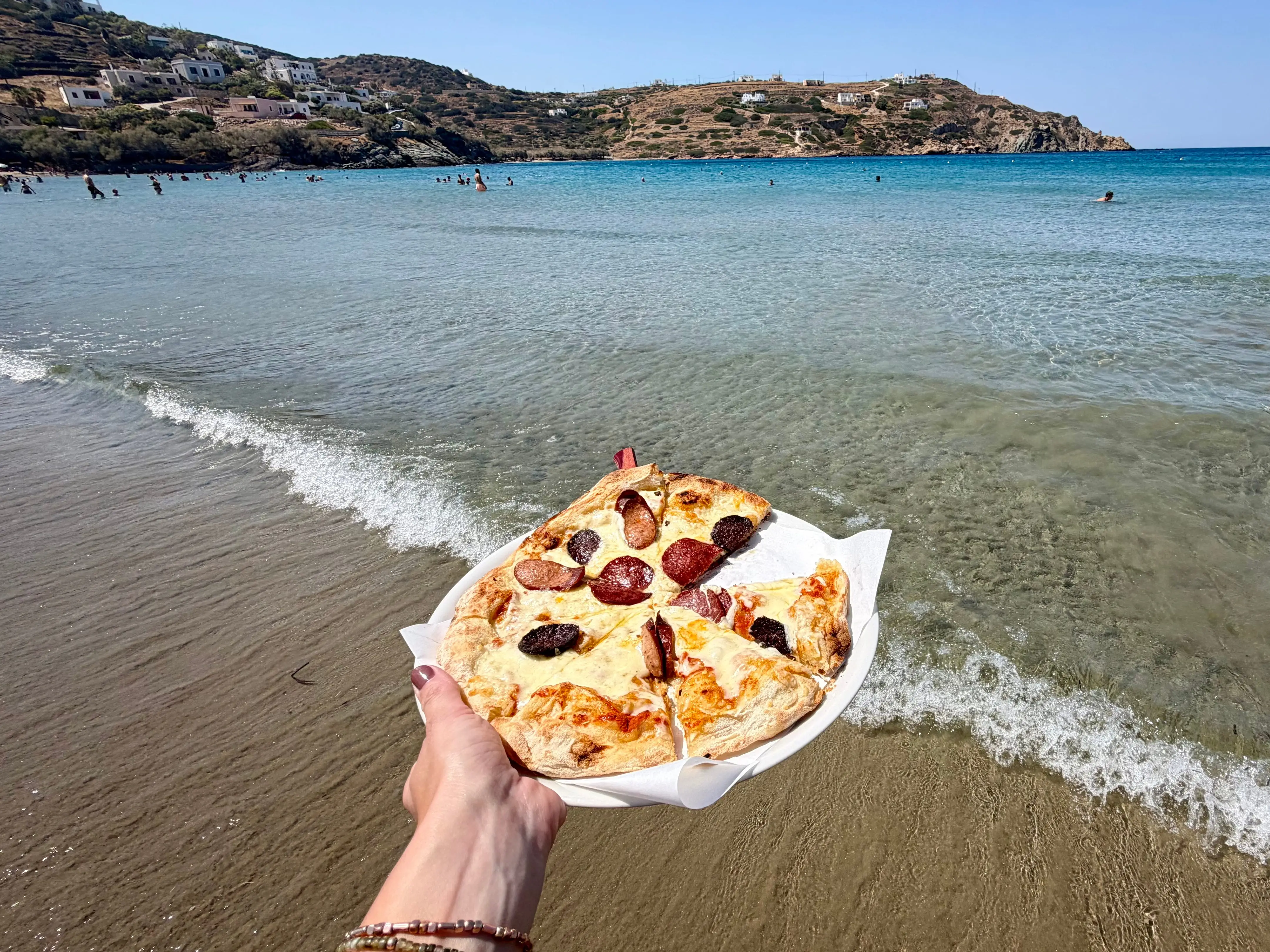 The writer is holding a pizza at the beach in Syros.