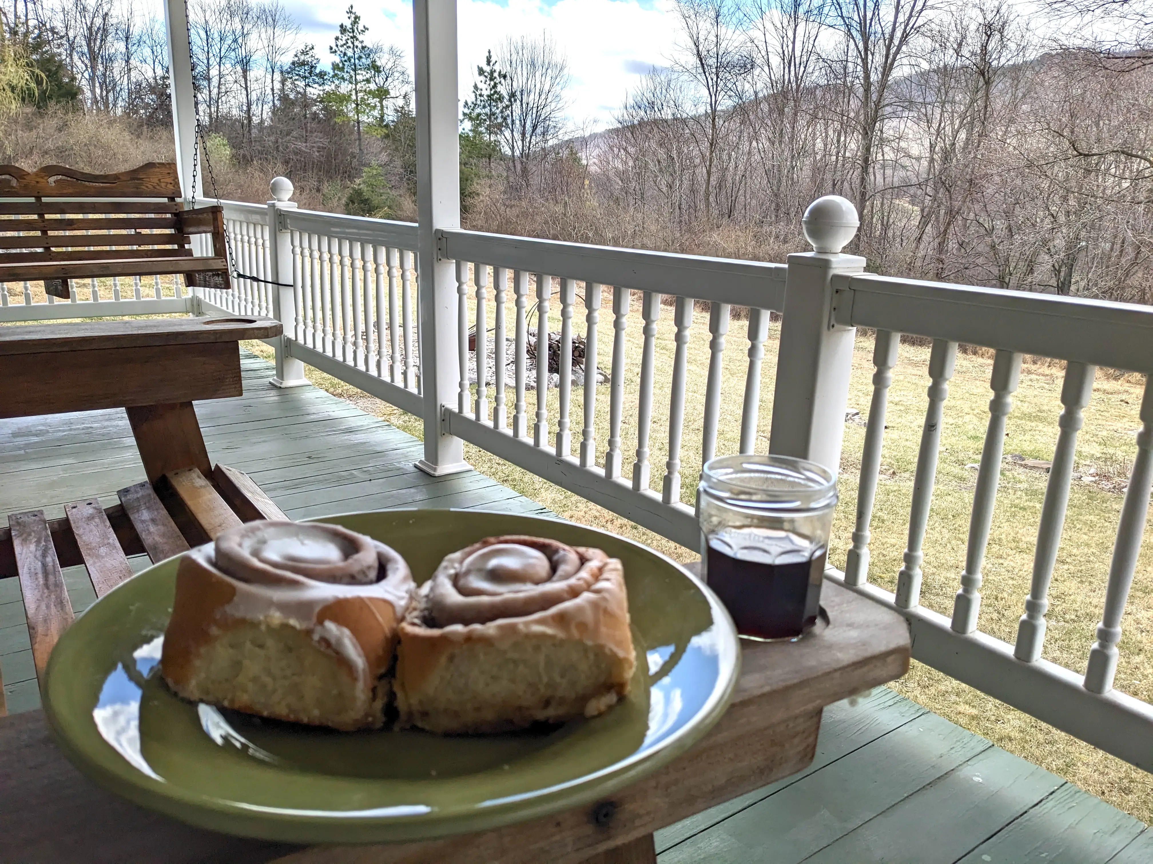 Two cinnamon buns sit on a green plate next to a drink on a porch overlooking mountains.