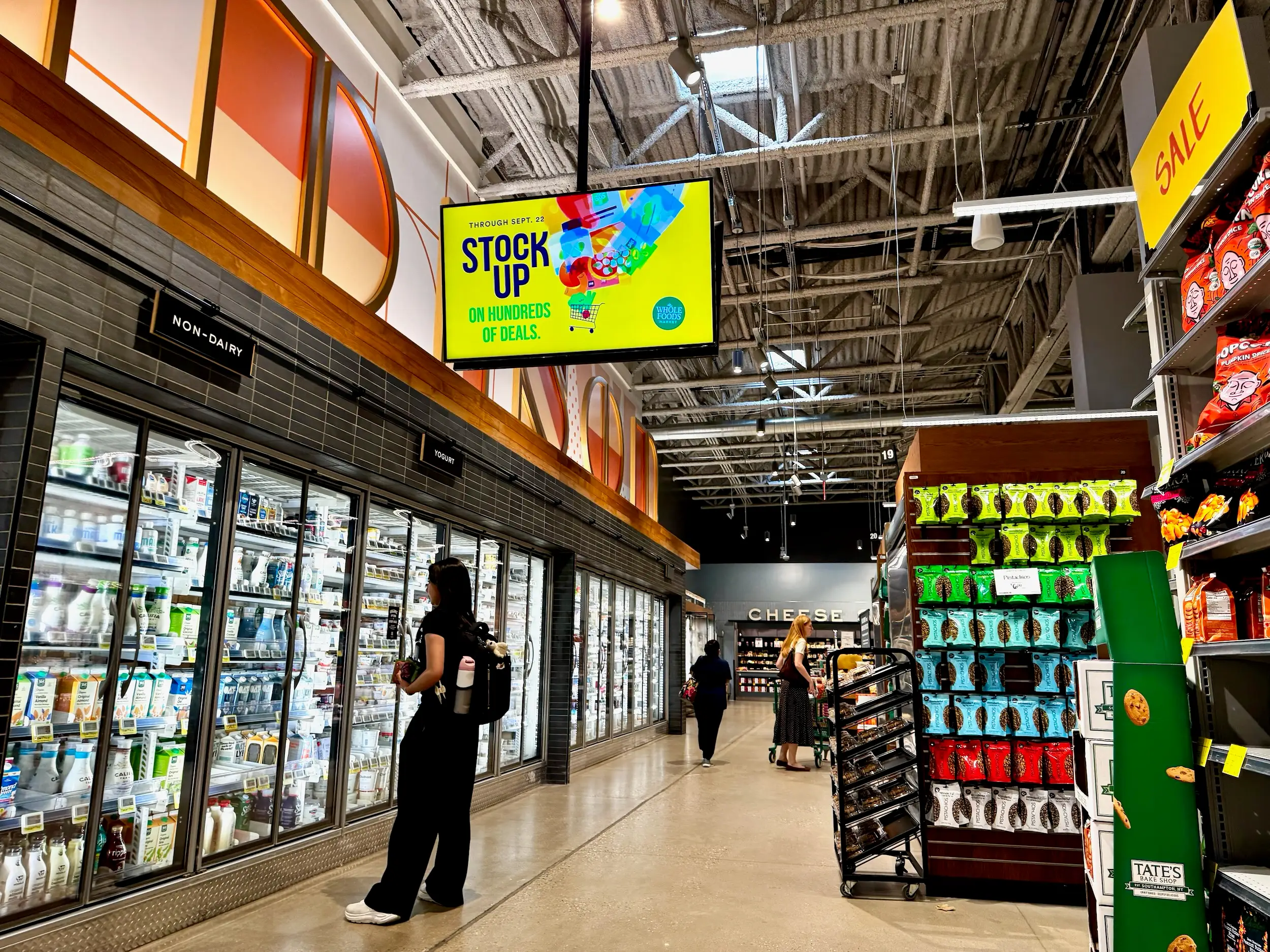 Shoppers browse refrigerated dairy cases and snack displays inside a Whole Foods Market aisle with sale signage overhead.