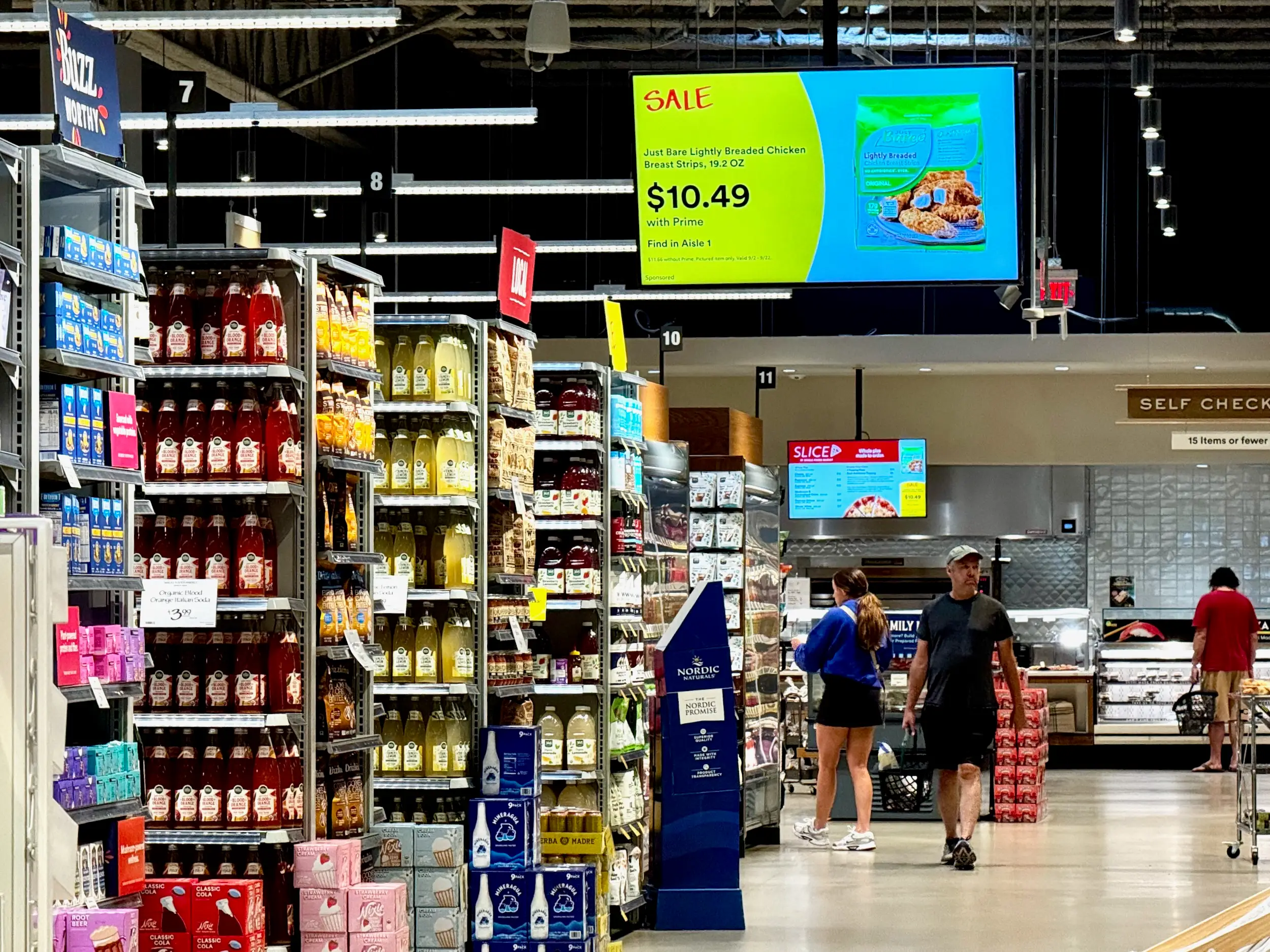 Shoppers walk through a grocery store aisle beneath a digital sale sign for Just Bare chicken strips.