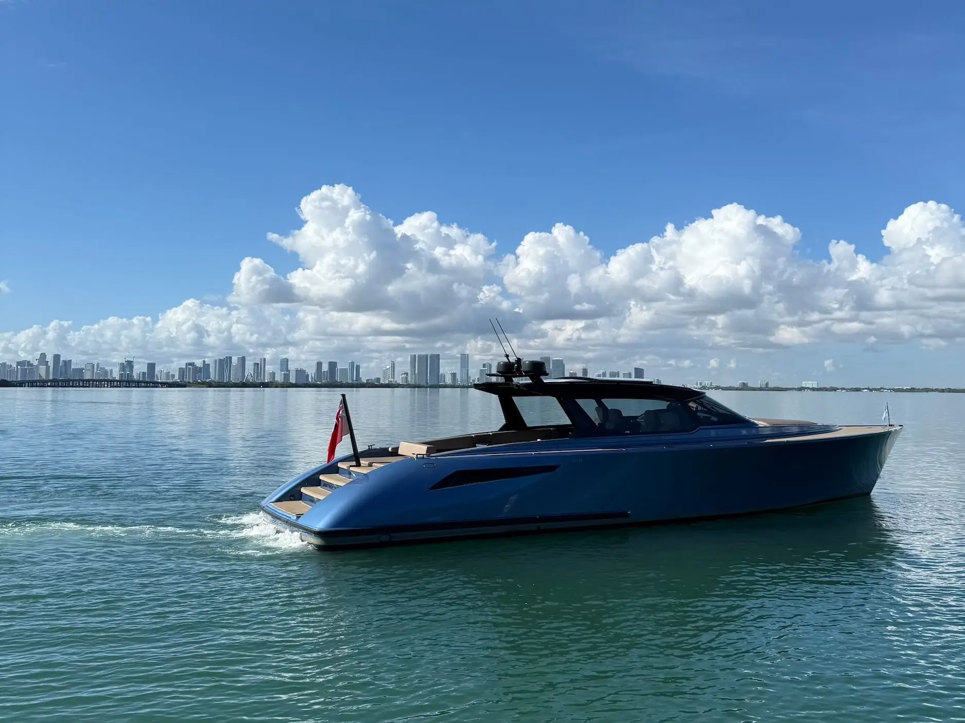 A sleek motor yacht cruises across calm turquoise water with a distant city skyline under a blue sky.