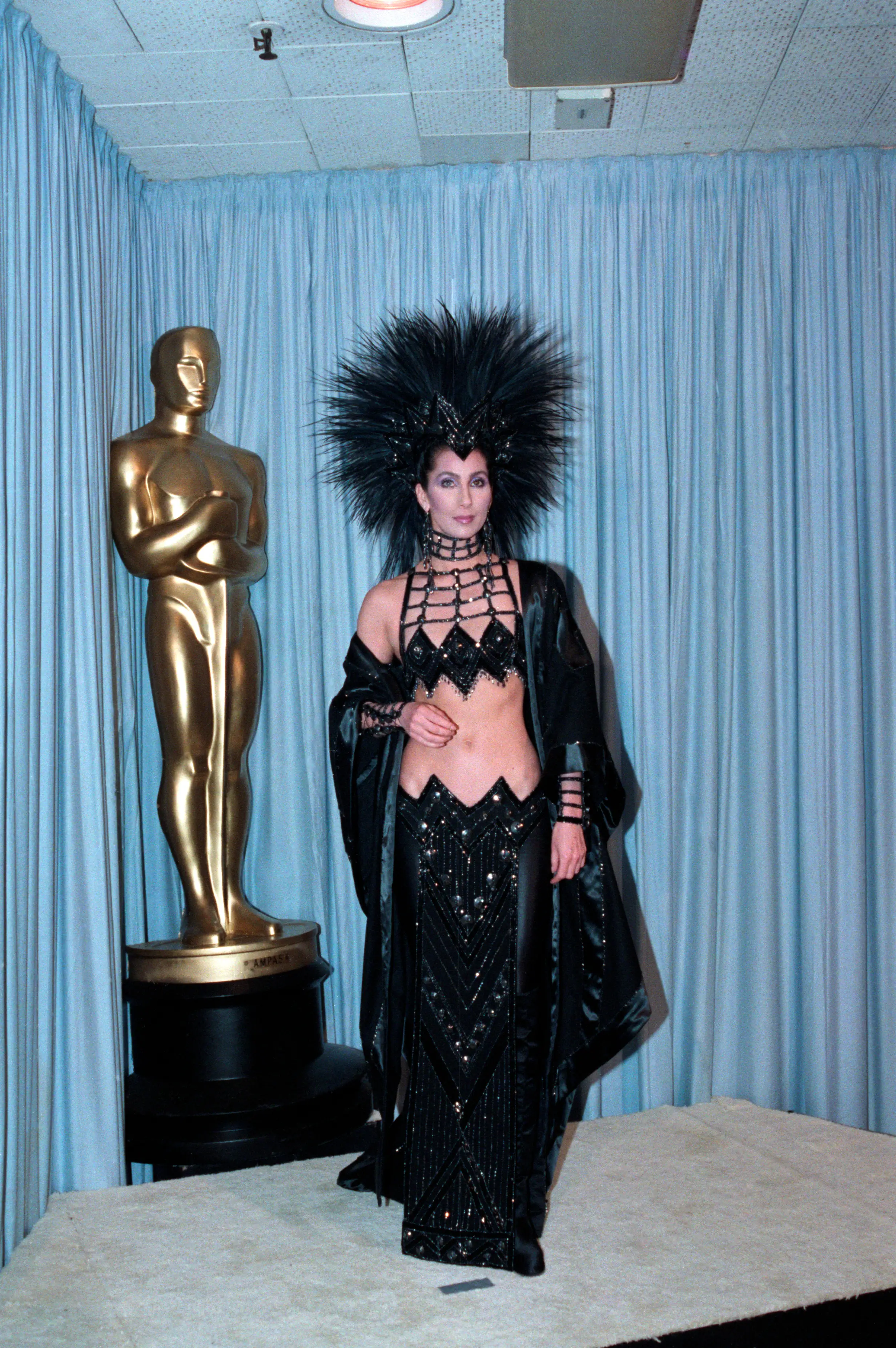 Cher stands in black sequined attire and spiked headpiece beside a large Oscar statuette.