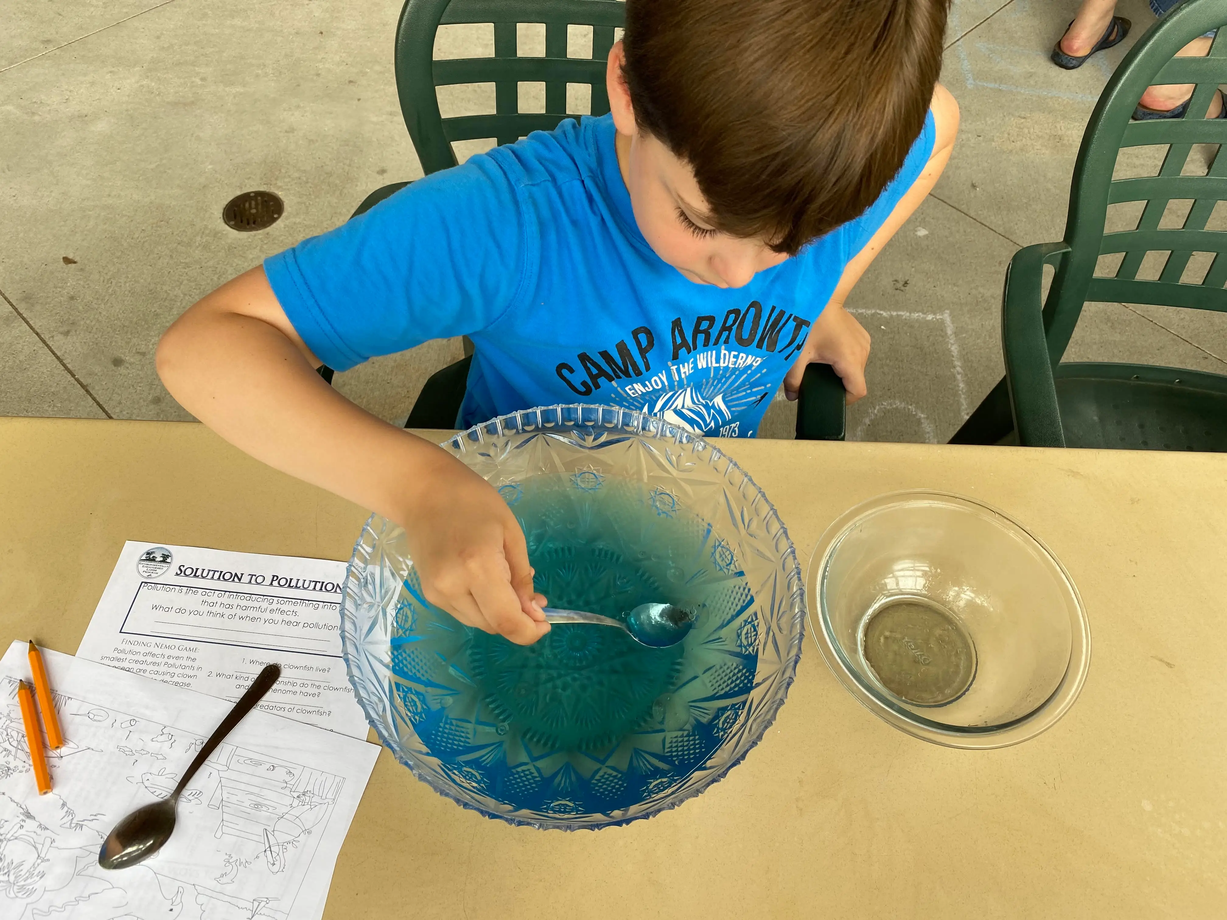 the author's son at a homeschooling class doing an experiment, he is holding a spoon in a bowl of blue liquid and there are papers on the table next to him