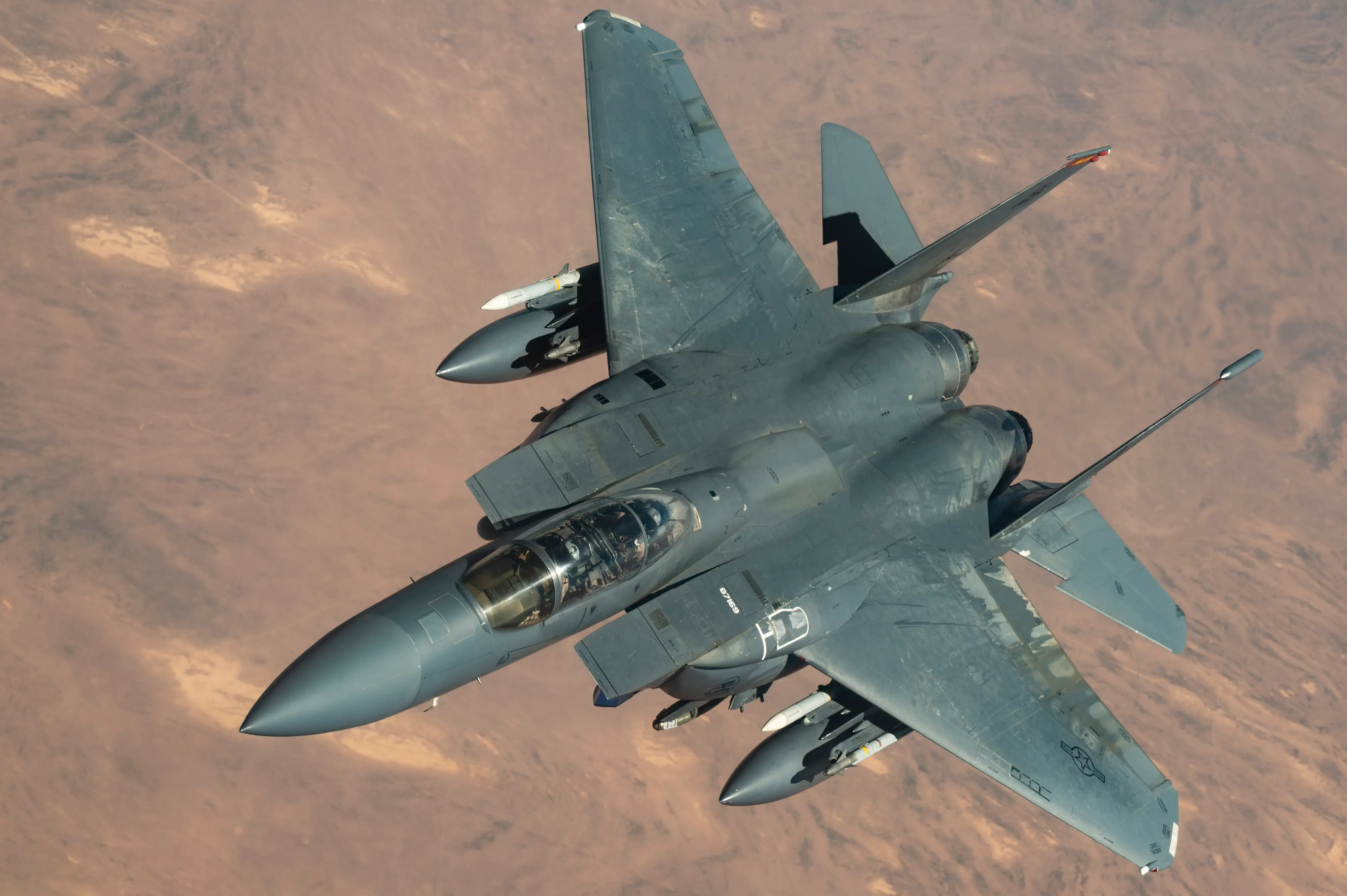 F-15E military fighter jet flies over a desert landscape with external tanks and visible underwing munitions.