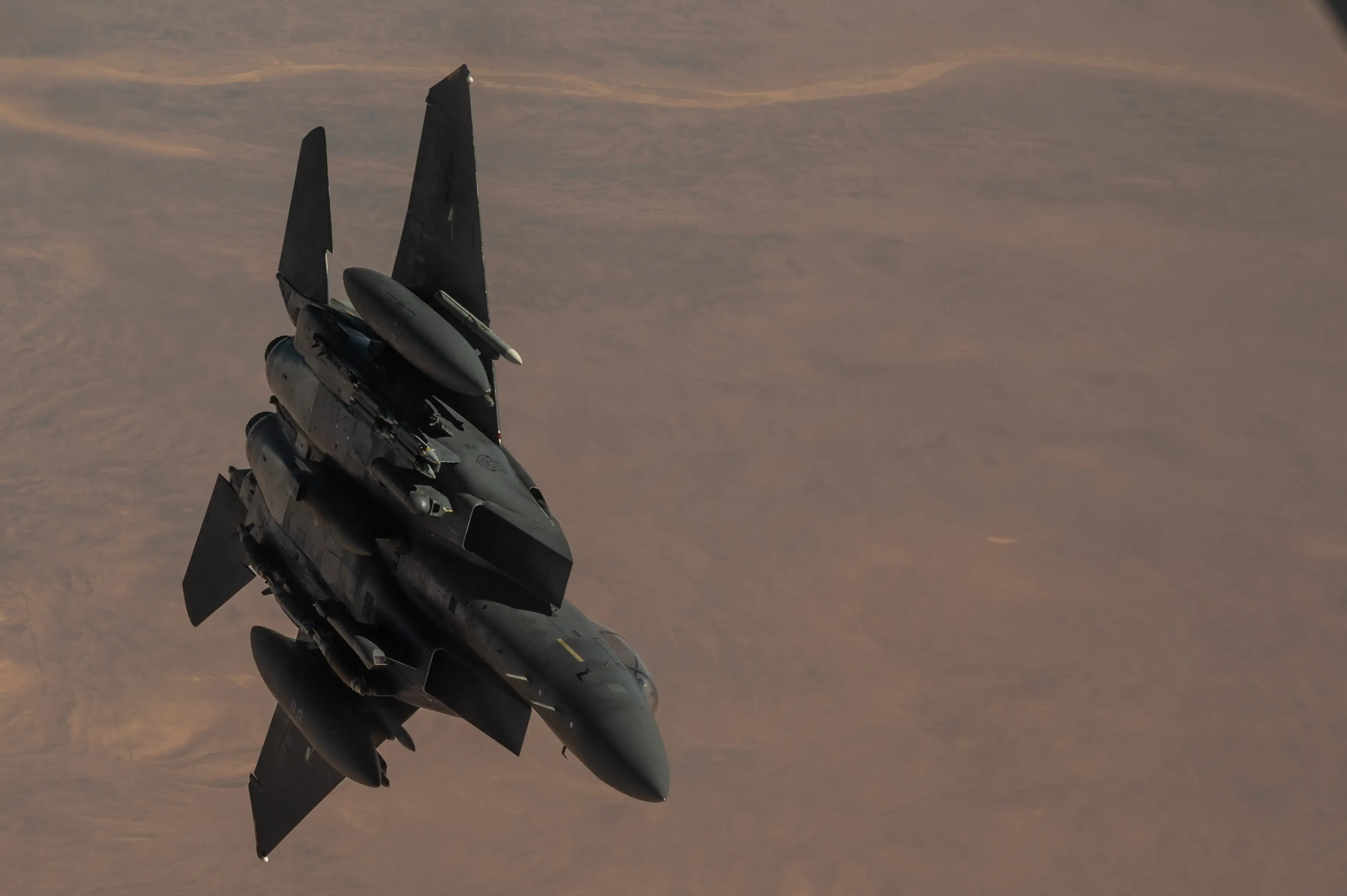 A fighter jet banks over a broad desert landscape in hazy light.