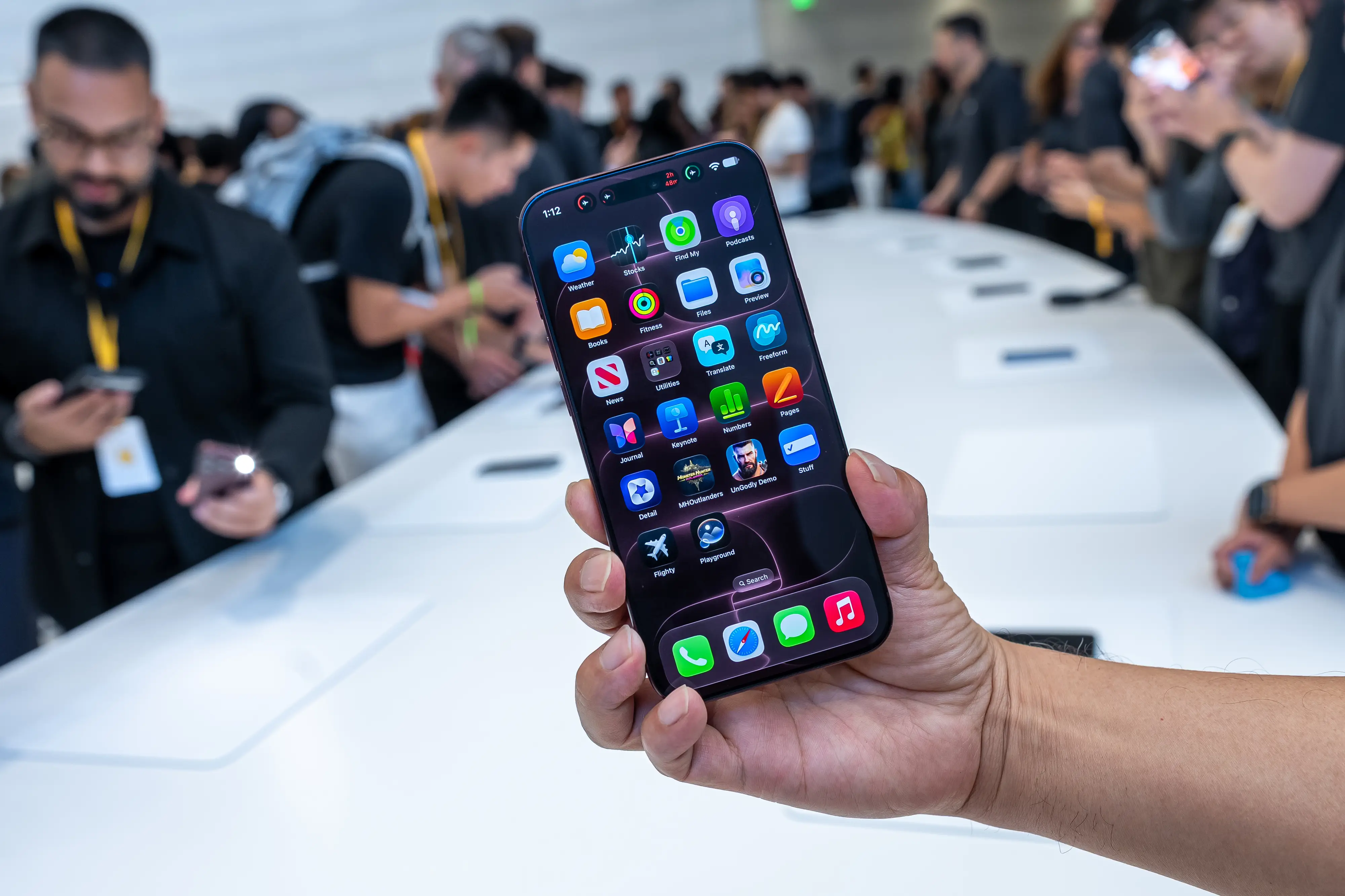 A hand holds an iPhone 18 Pro at a long white table.