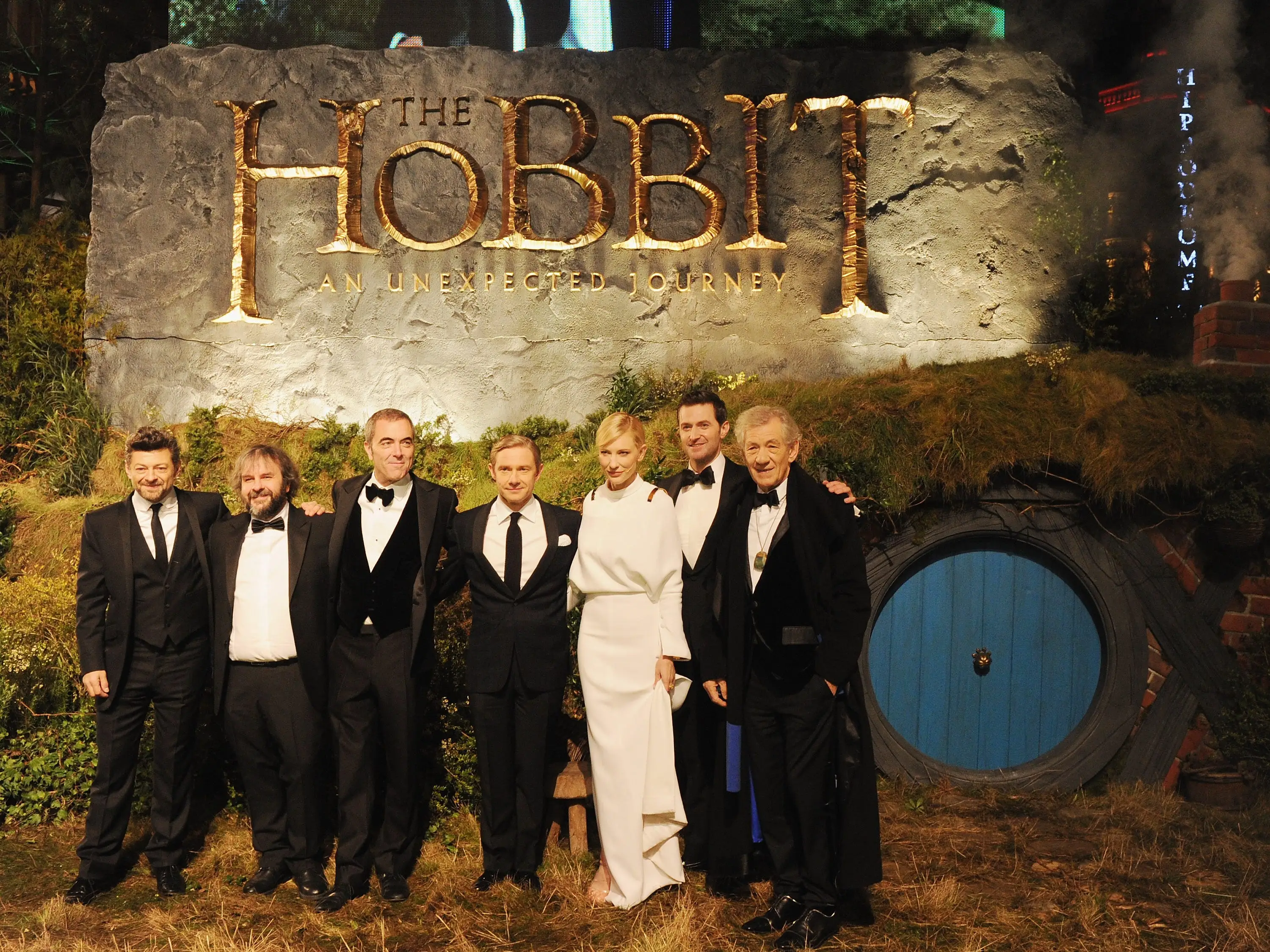 Several actors in tuxedos and an actress in a white dress pose at a movie premiere.