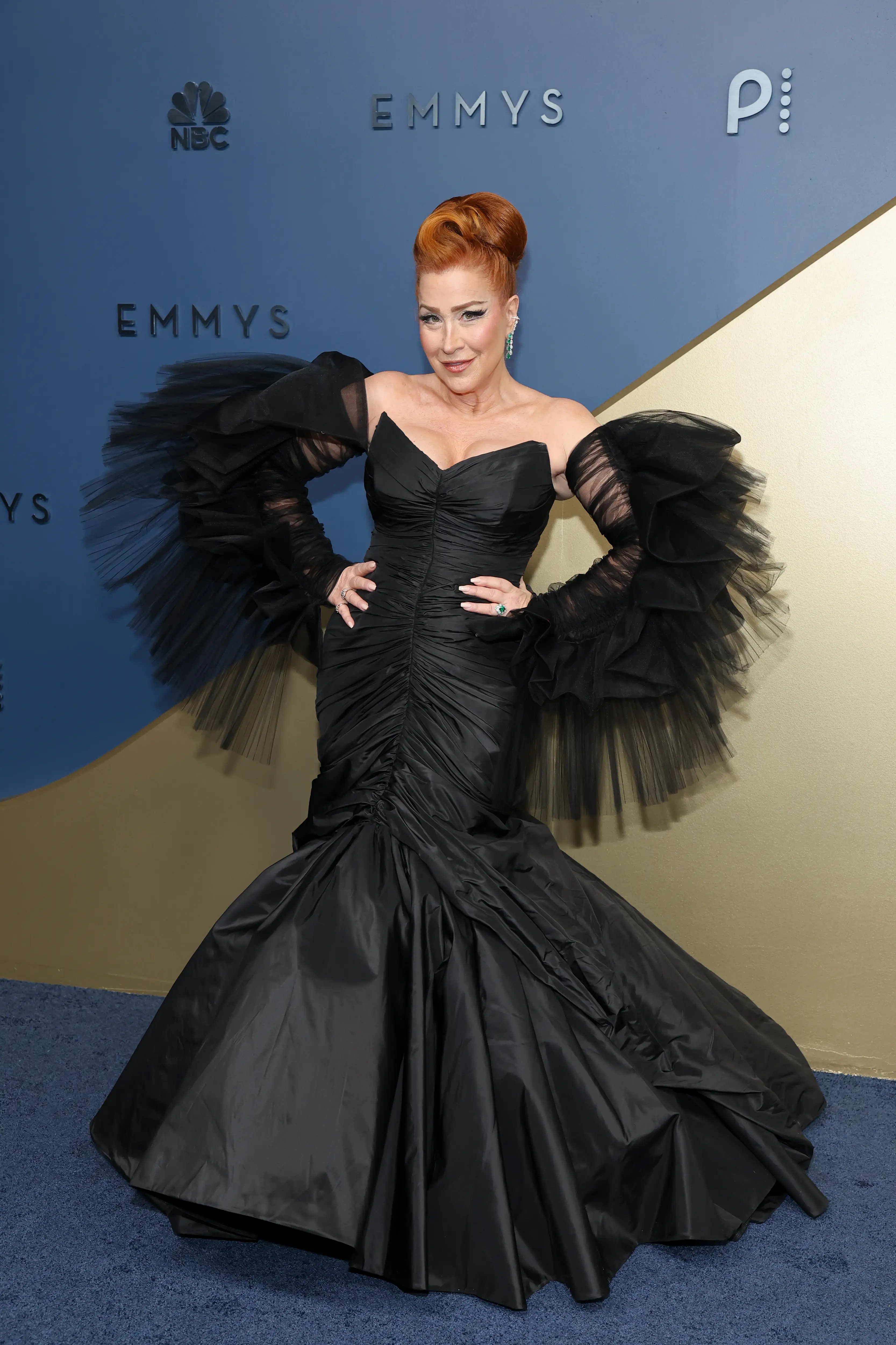 Lisa Ann Walter poses in a dramatic black gown before a blue and gold Emmys backdrop.