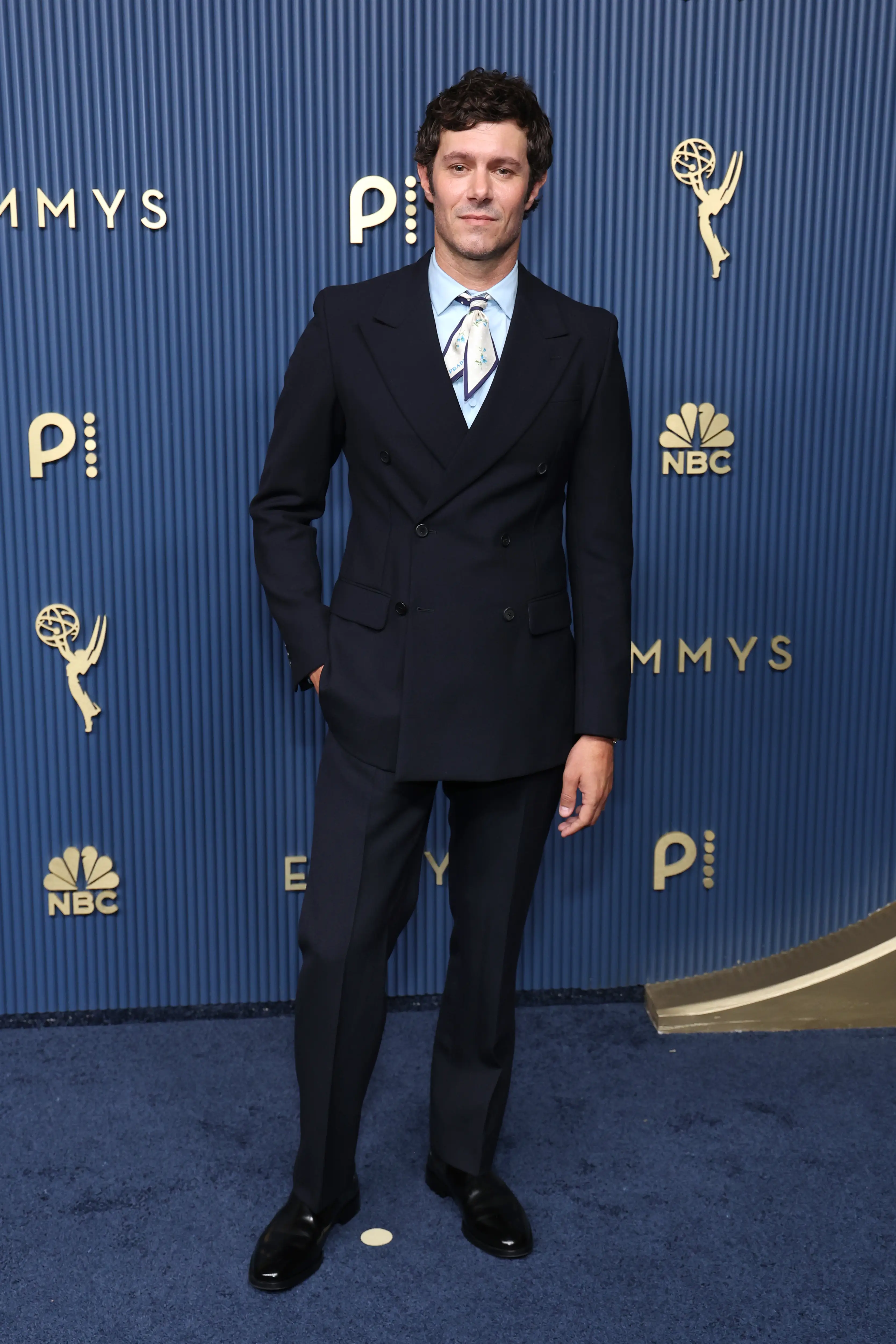 Adam Brody stands on a blue carpet in a navy suit before an Emmys and NBC step-and-repeat backdrop.