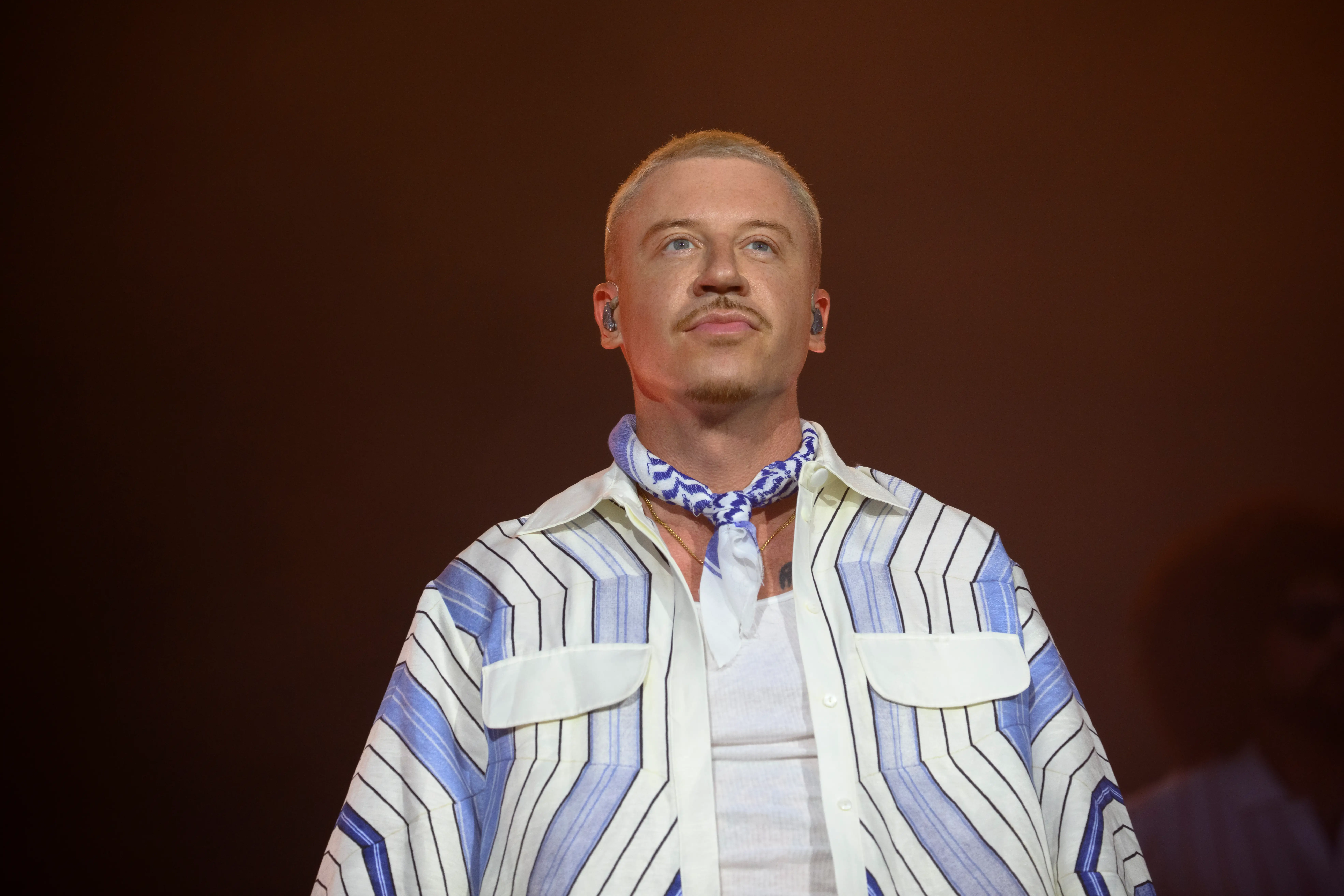 Macklemore performs onstage wearing a blue-and-white striped shirt and neck scarf against a dark warm-lit background.