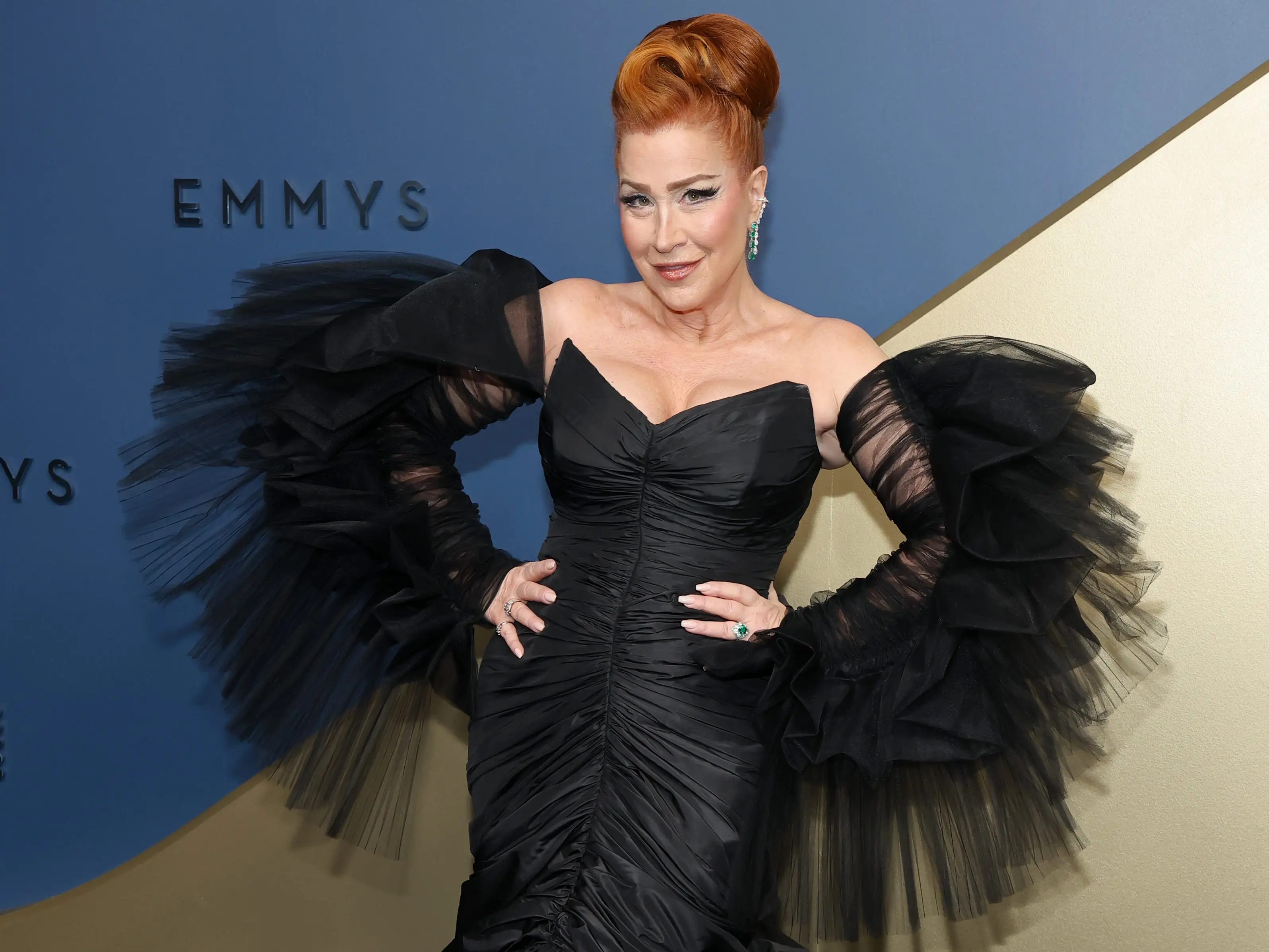 Lisa Ann Walter poses in a dramatic black gown before a blue and gold Emmys backdrop.