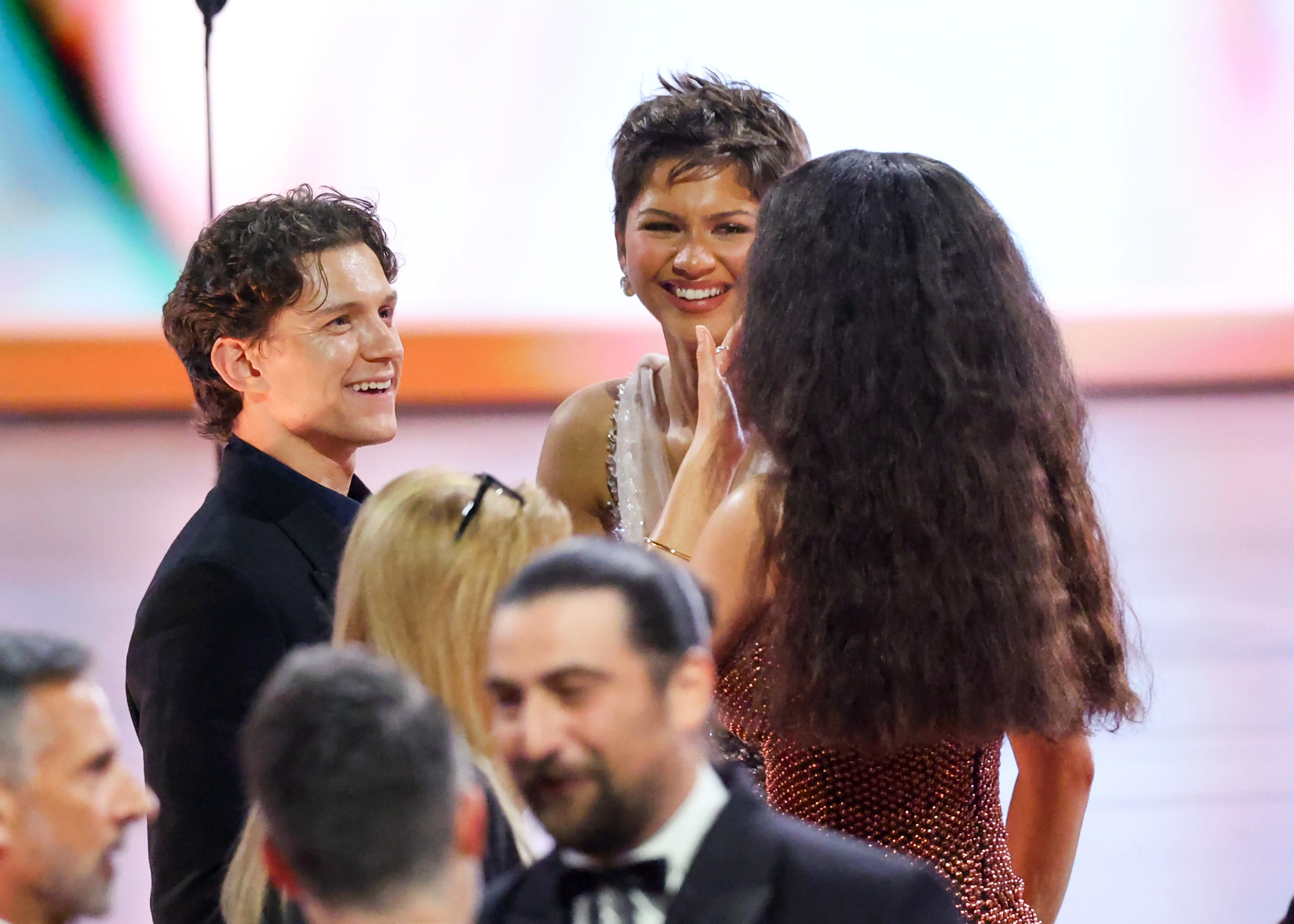 Tom Holland smiles while speaking with Zendaya and other attendees at a formal indoor event.