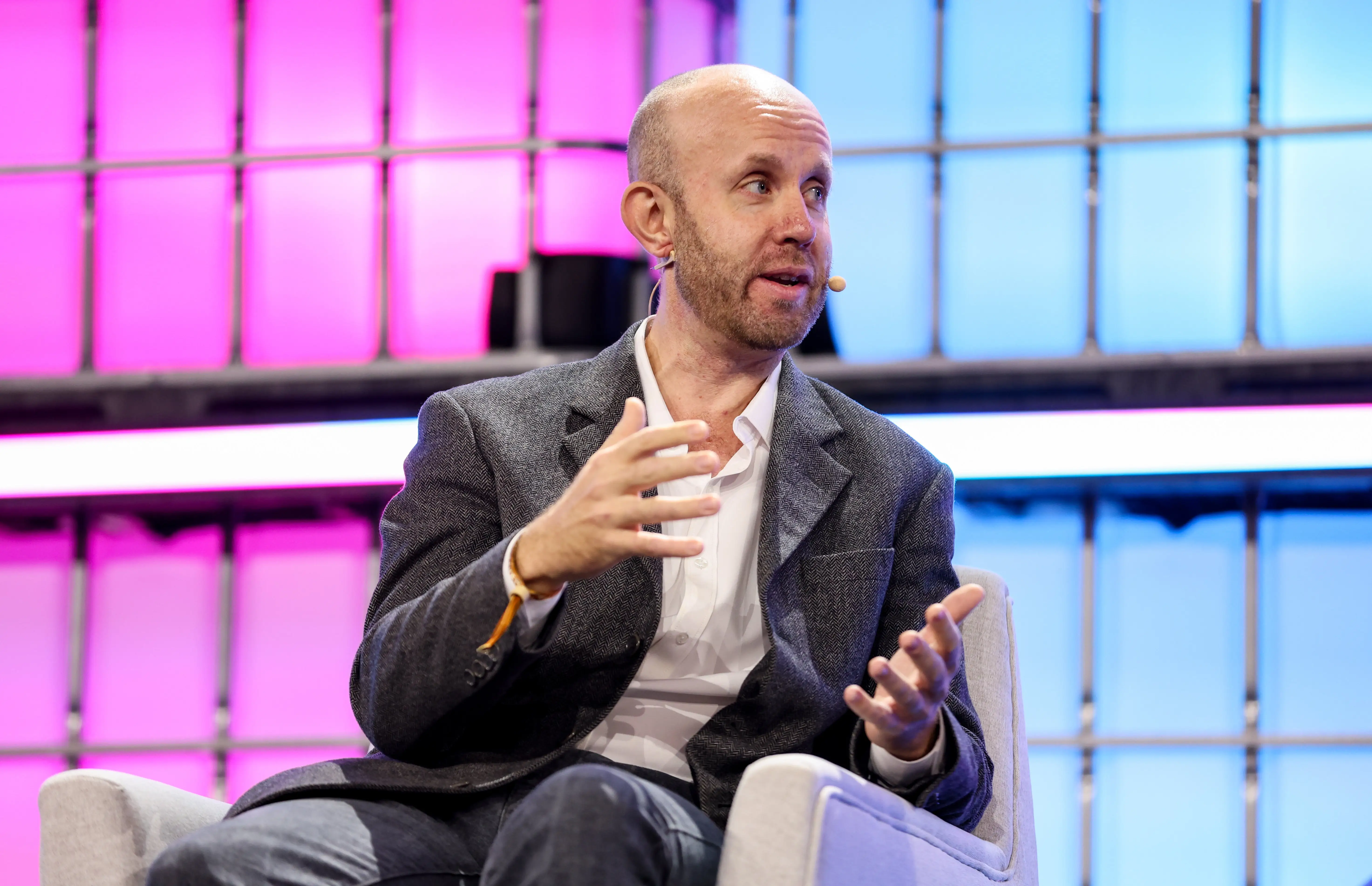 Bald speaker in a blazer gestures while seated onstage before a pink and blue illuminated backdrop.