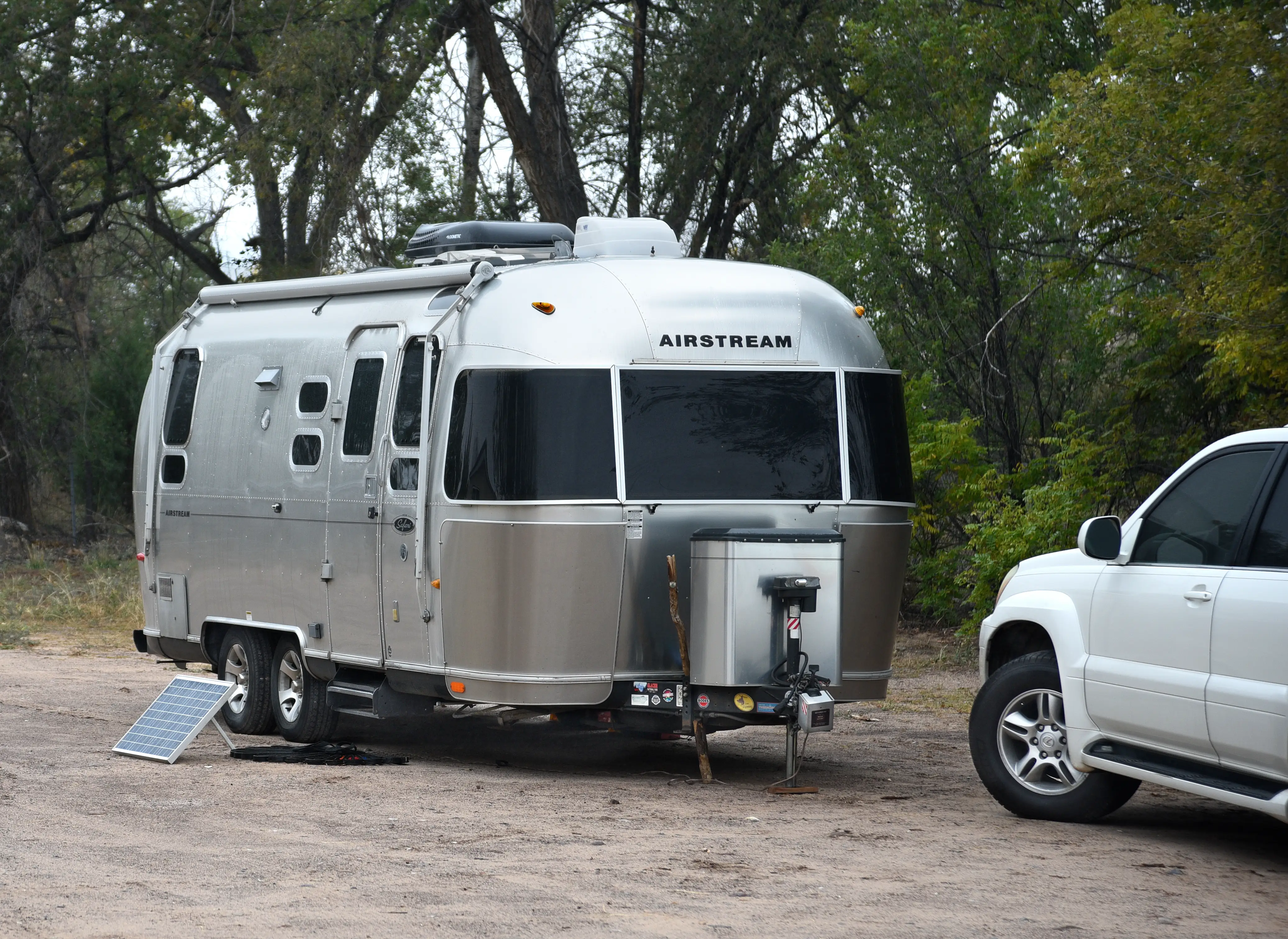 Silver AIRSTREAM travel trailer parked on a dirt campsite beside a white SUV and trees.