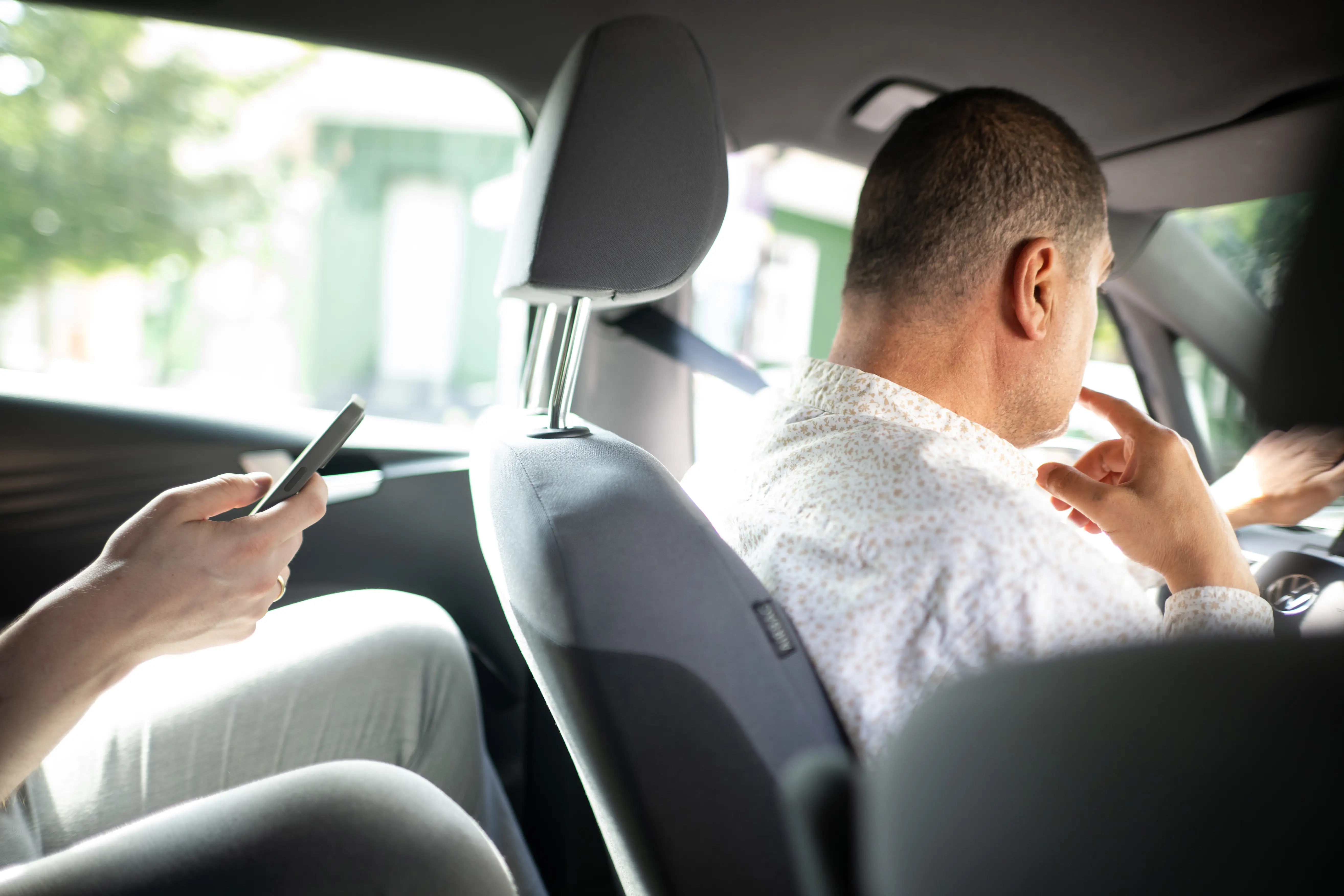 Passenger uses a smartphone while another person sits at the steering wheel inside a car.