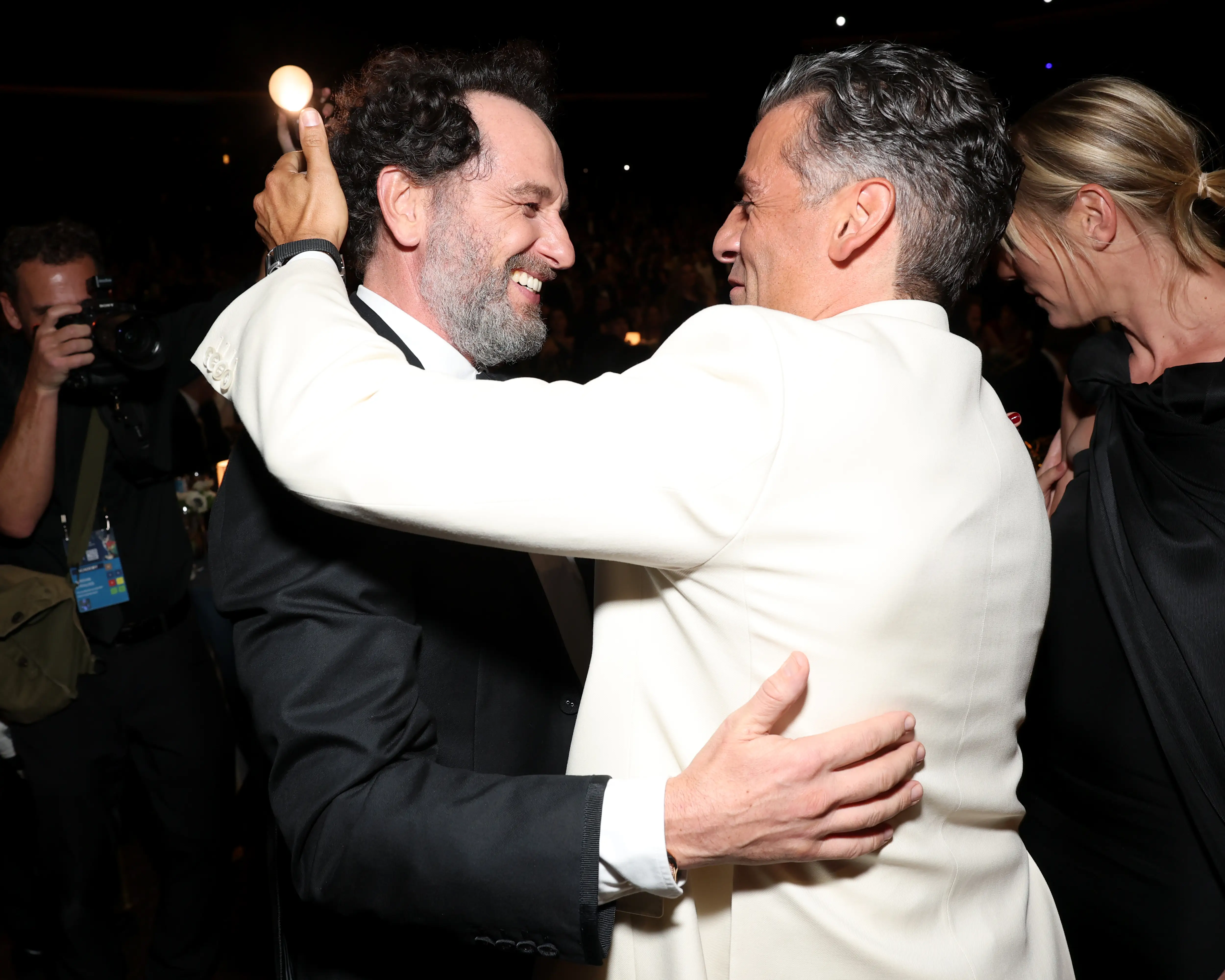 Matthew Rhys and Oscar Isaac embrace at a formal event while photographers and attendees stand nearby.