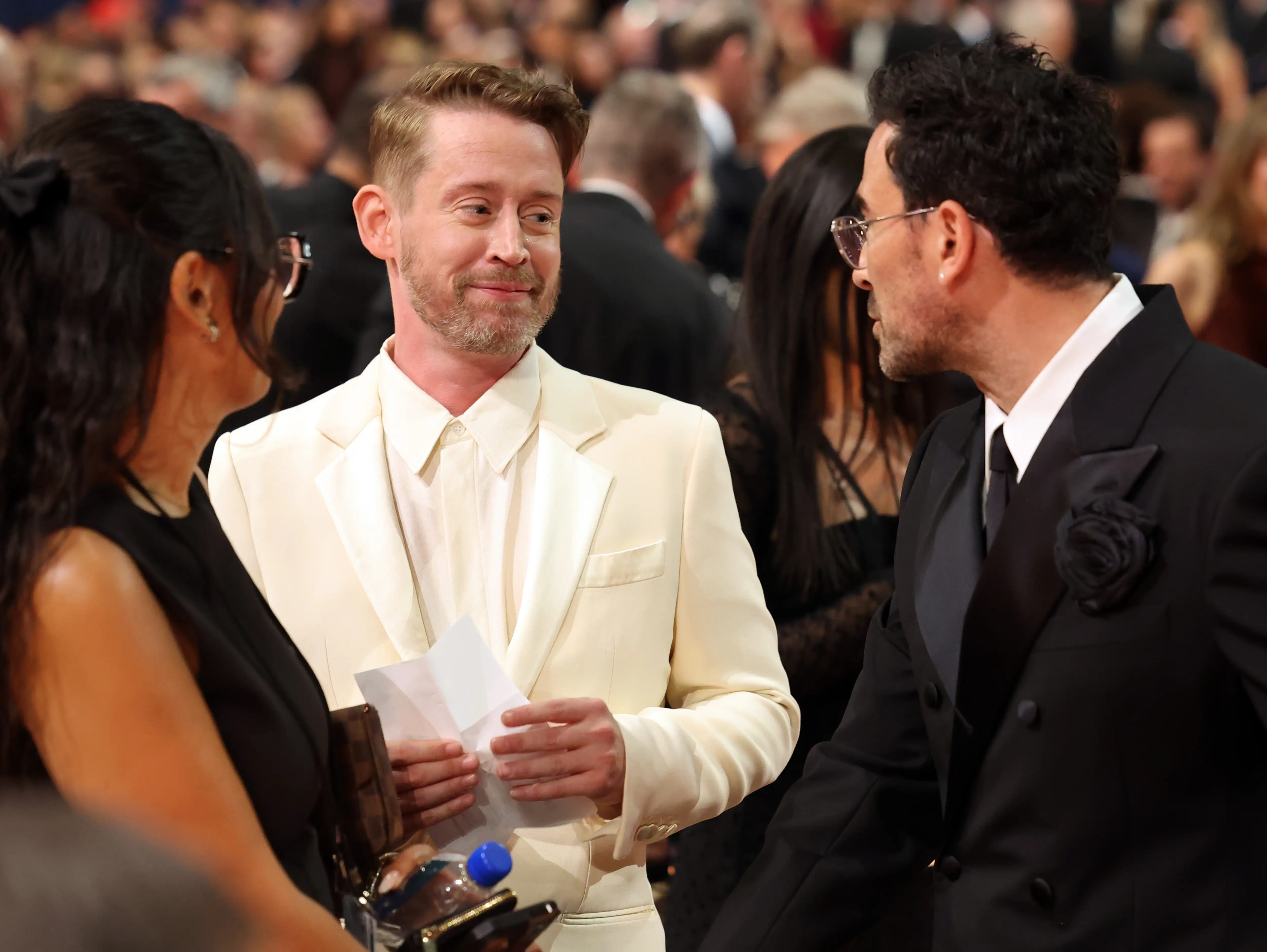 Macaulay Culkin speaks with Dan Levy in formalwear amid a crowded red carpet event.