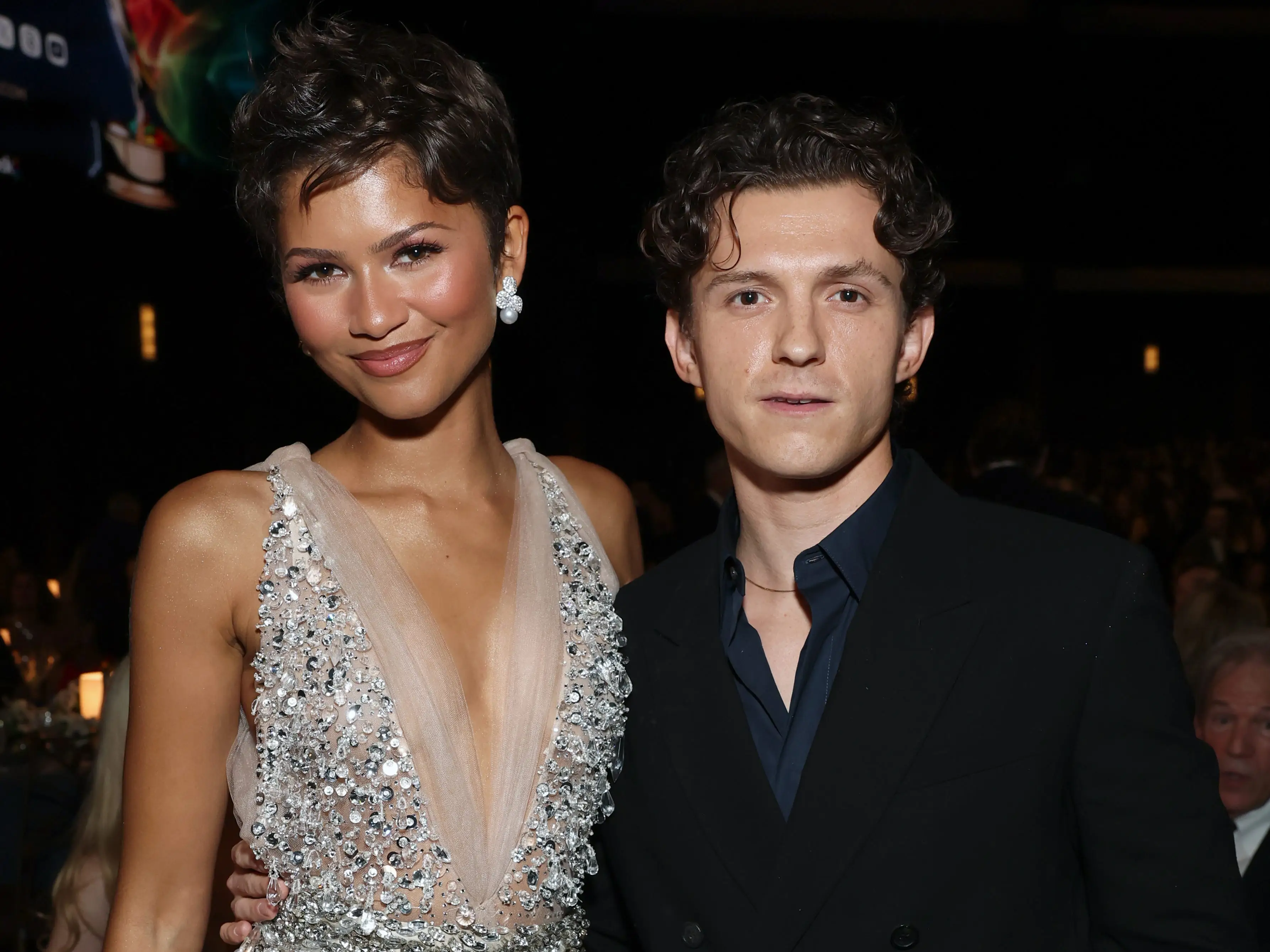 Zendaya and Tom Holland pose together in formal attire at a dimly lit indoor event.