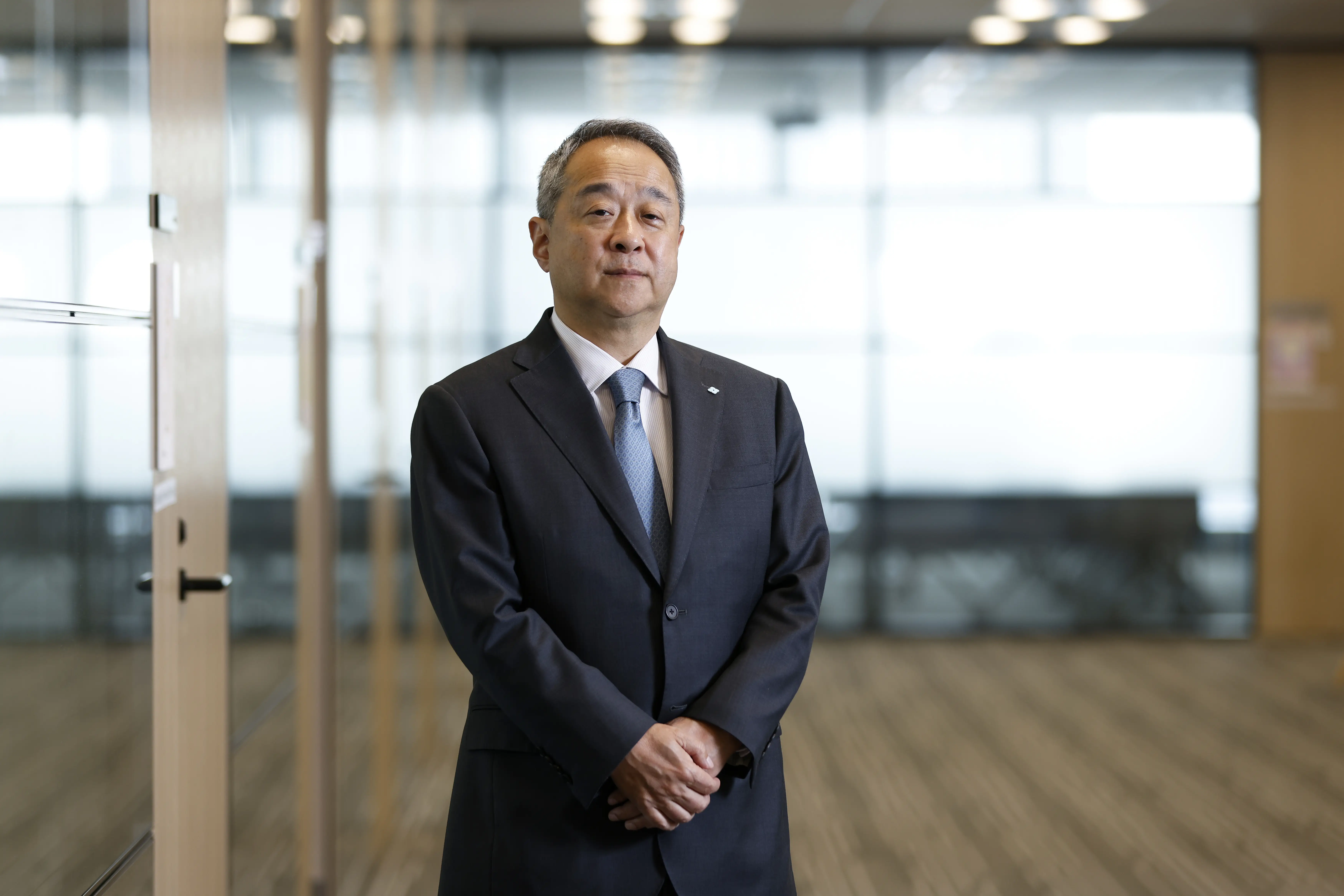 A suited executive stands with hands clasped in a modern glass-walled office.