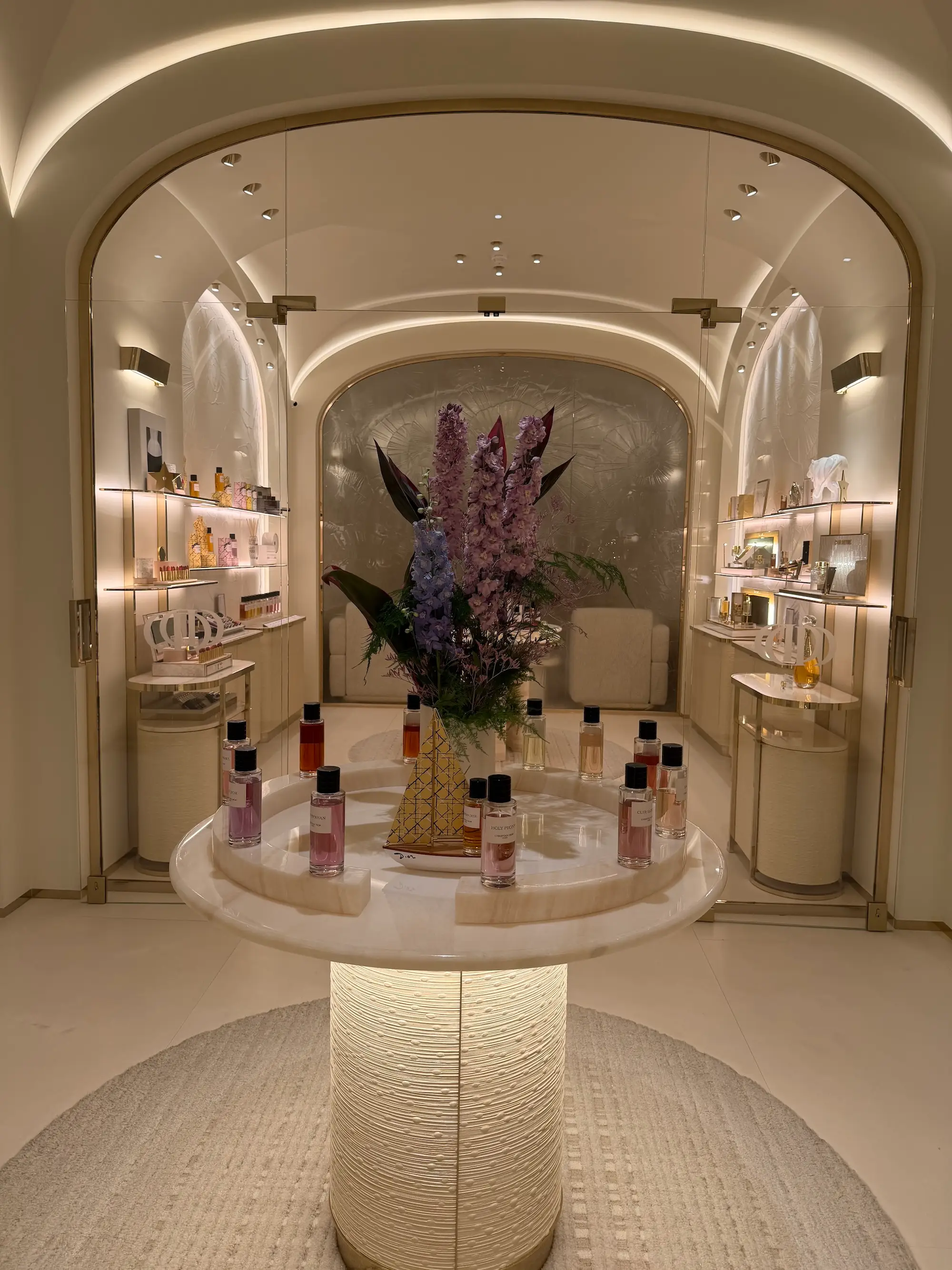 Illuminated luxury cosmetics boutique displays perfume bottles around a flower arrangement on a central round table.
