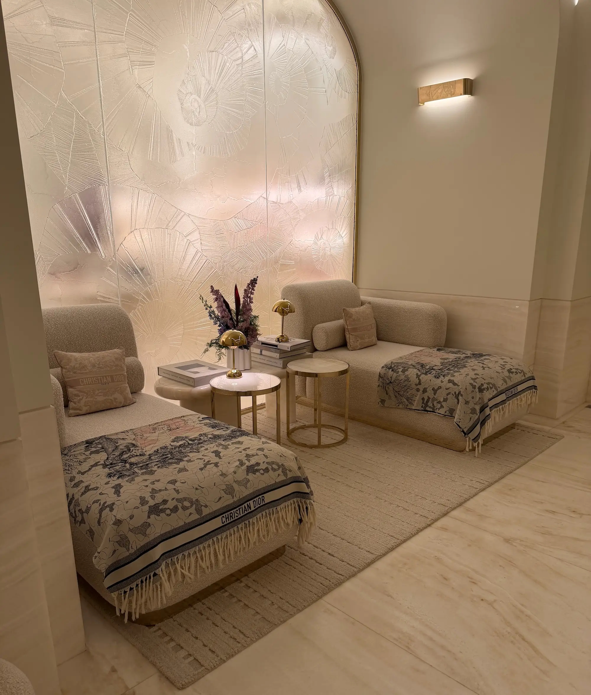 Elegant lounge alcove with two upholstered chaise seats, Christian Dior throws, brass tables, flowers, and illuminated textured wall art.