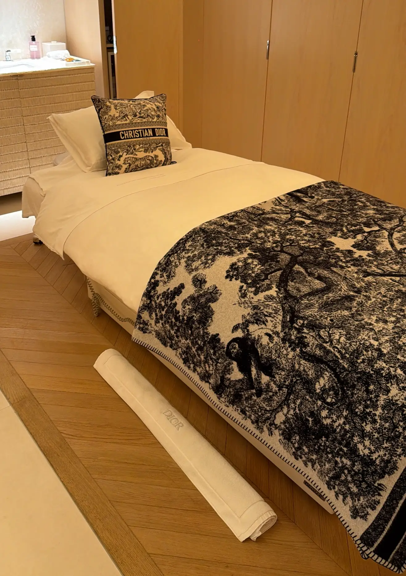 Massage bed with a Christian Dior pillow and a patterned blanket beside wood wardrobes in a warmly lit room.