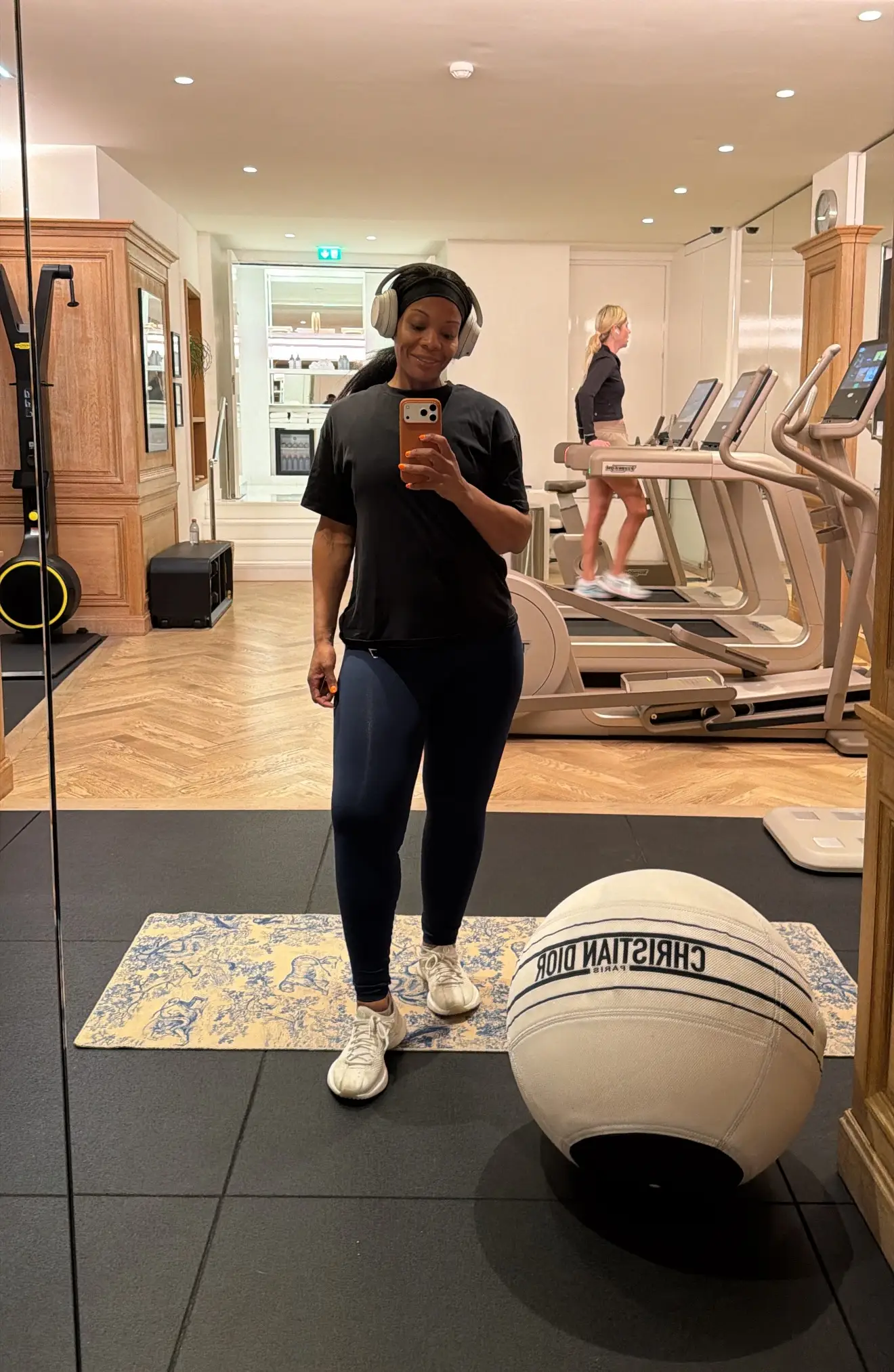 Person takes a mirror selfie in a gym with treadmills and a CHRISTIAN DIOR exercise ball.