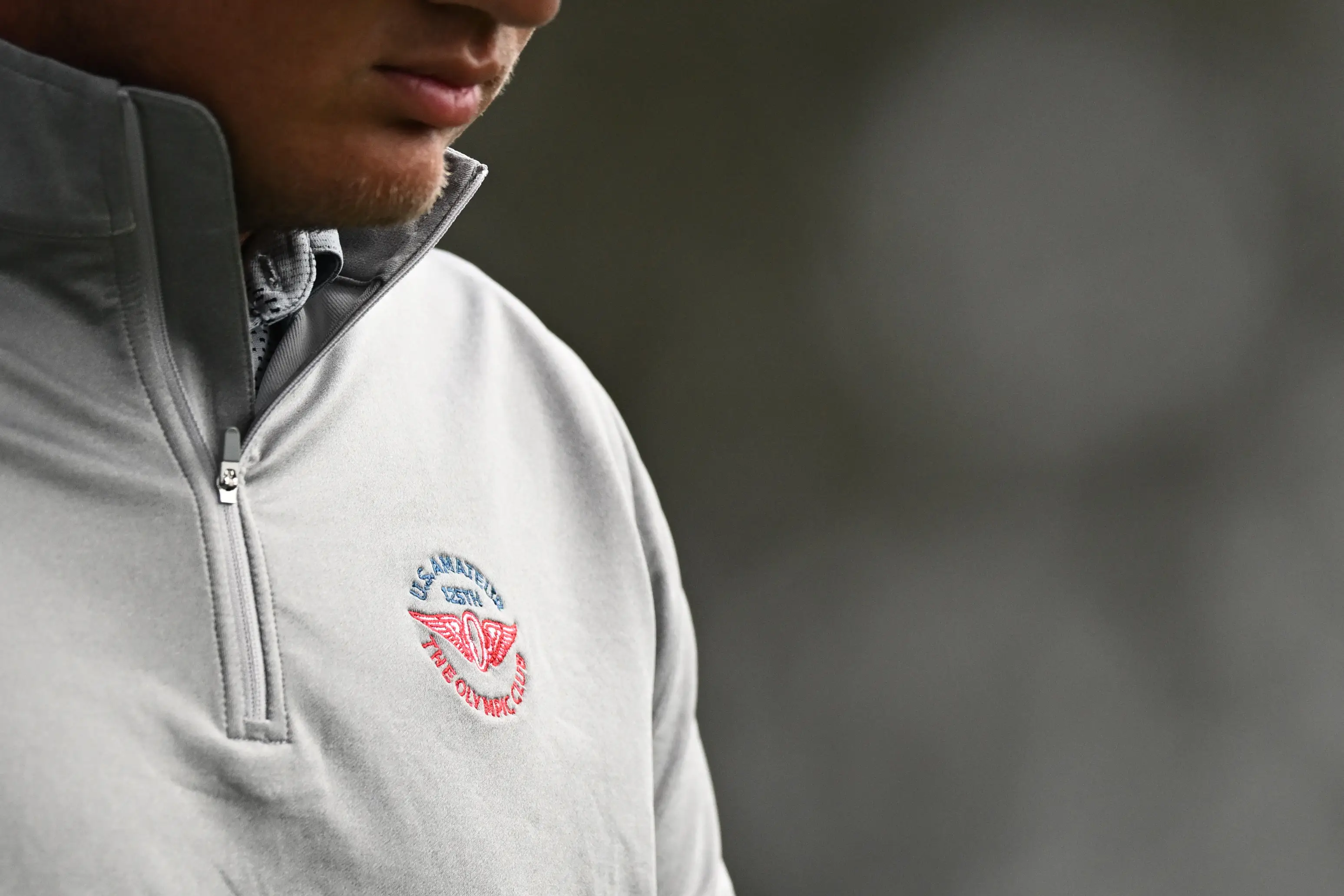 Cropped close-up of a person wearing a gray quarter-zip pullover with an embroidered chest logo.