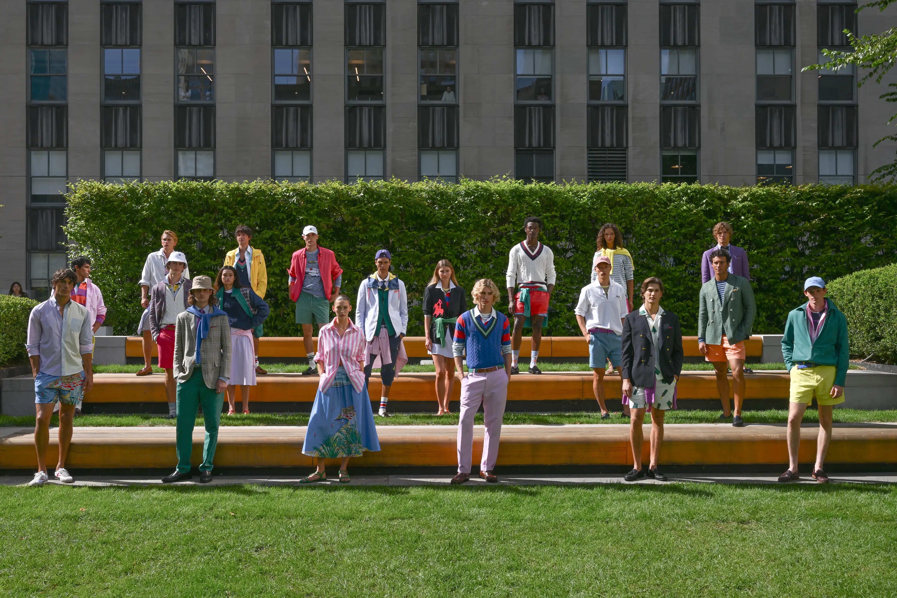Models in colorful preppy outfits stand spaced on tiered benches in a landscaped courtyard before a tall building.