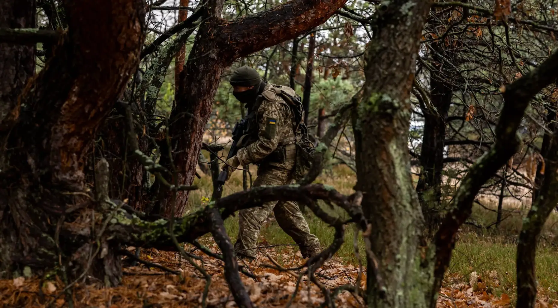 Camouflaged soldier with a Ukrainian flag patch carries a rifle through a dense wooded area.