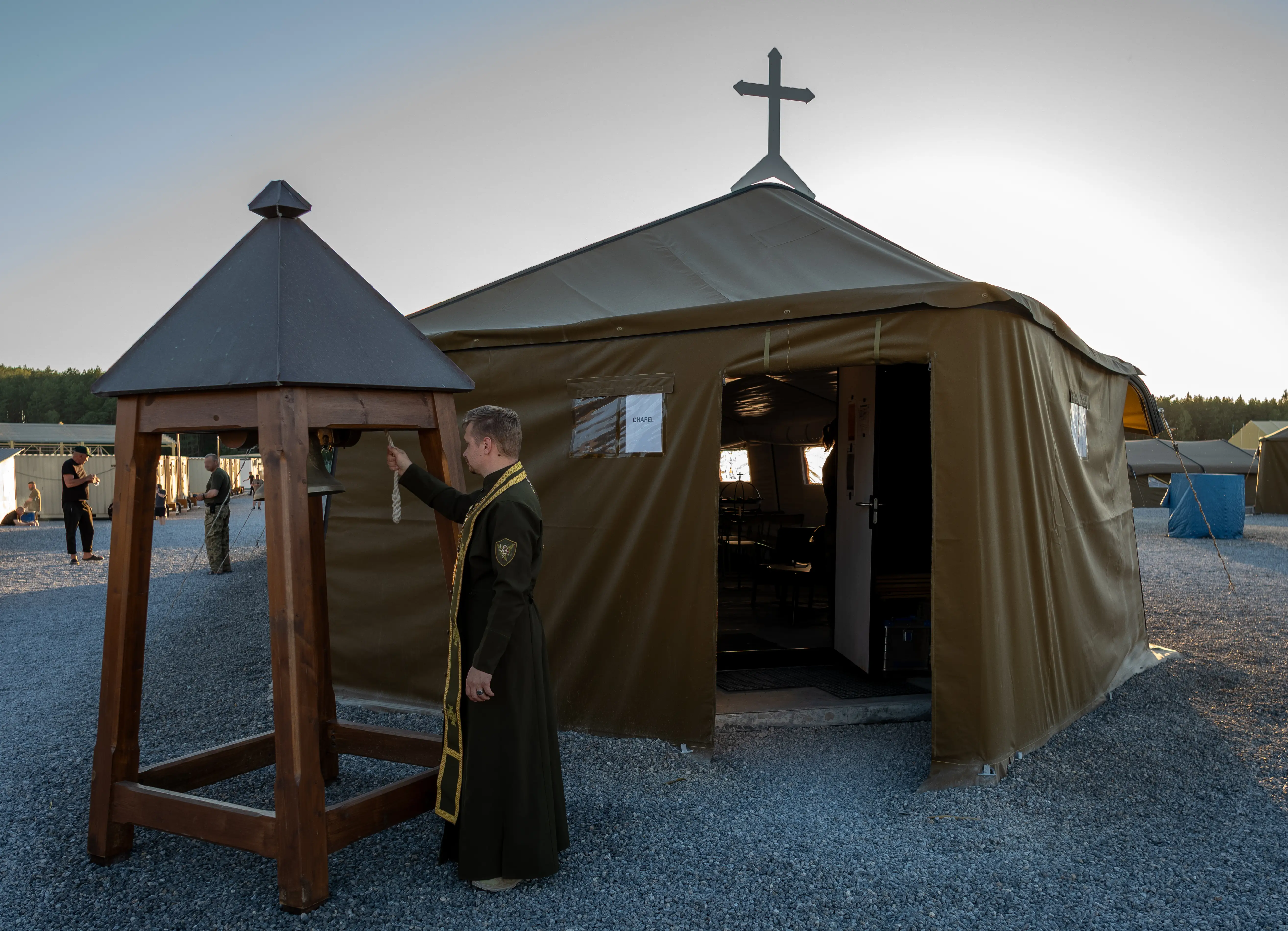Cleric in dark robes rings a bell beside a tent chapel with a cross at an outdoor camp.