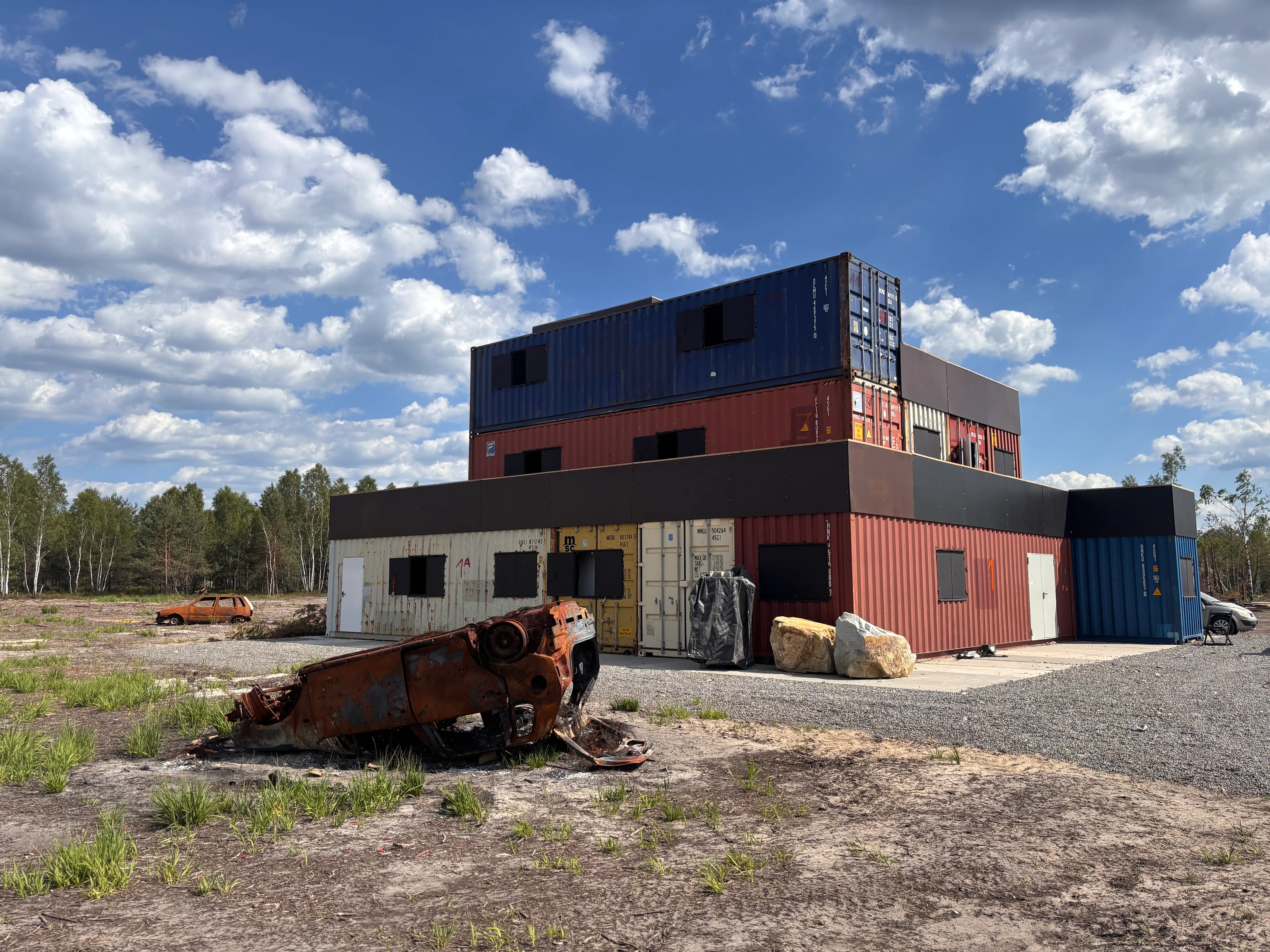 Stacked shipping containers form a multi-level structure beside rusted vehicle wrecks under a cloudy blue sky.