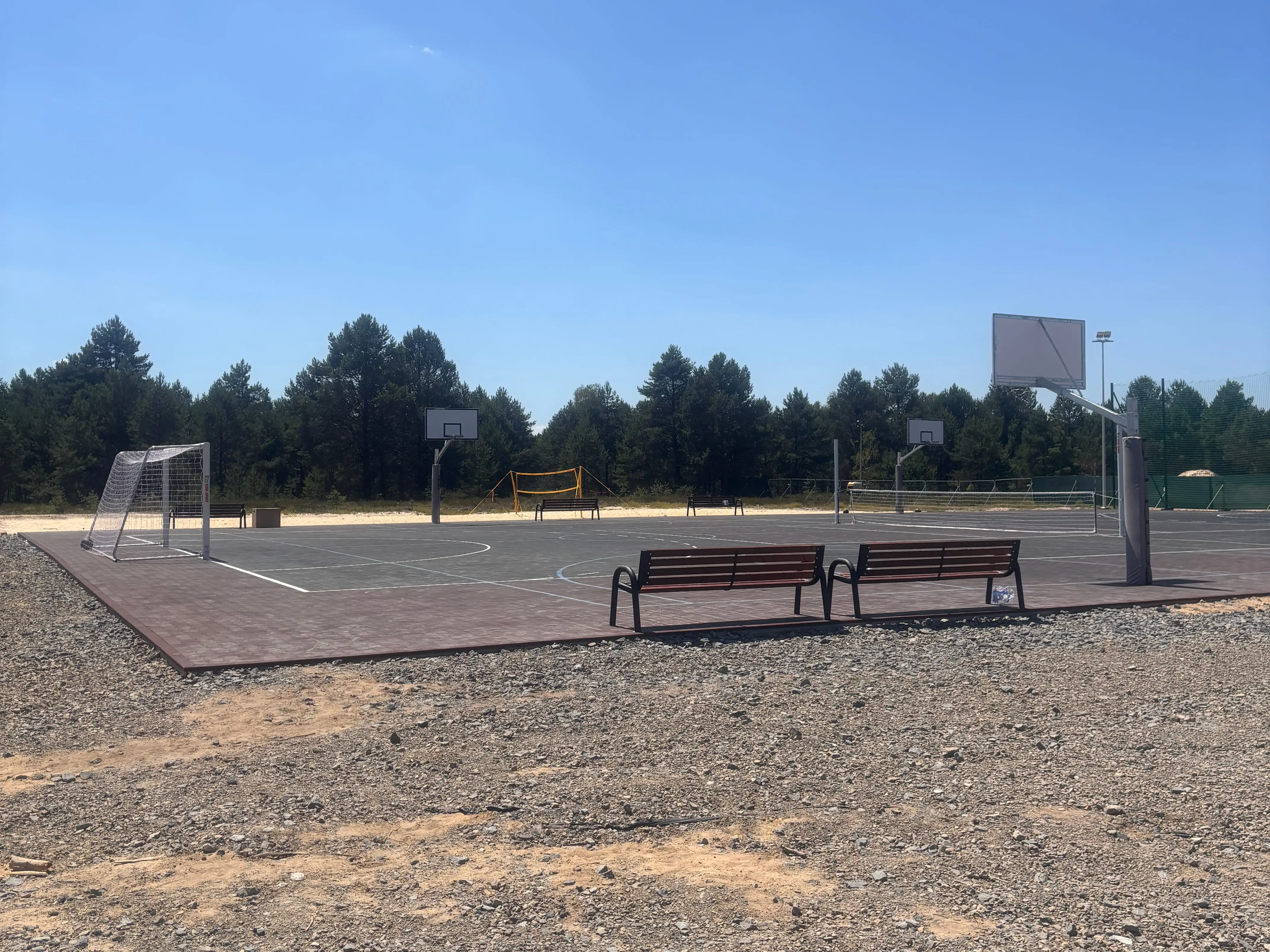 Empty outdoor sports court with basketball hoops, a soccer goal, benches, trees, and clear blue sky.