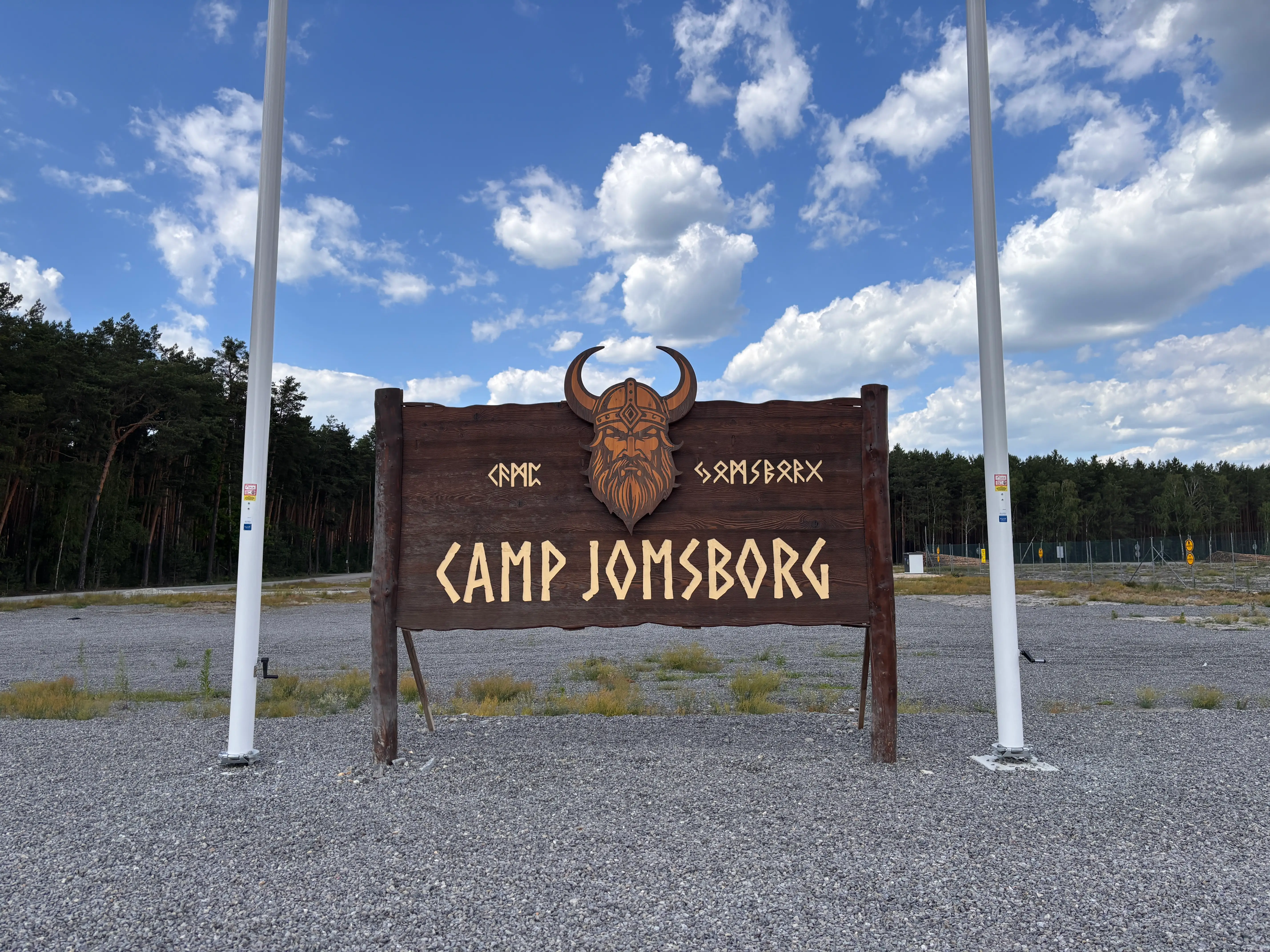 Wooden CAMP JOMSBORG sign with a Viking-style emblem stands between flagpoles beside a forested gravel area.