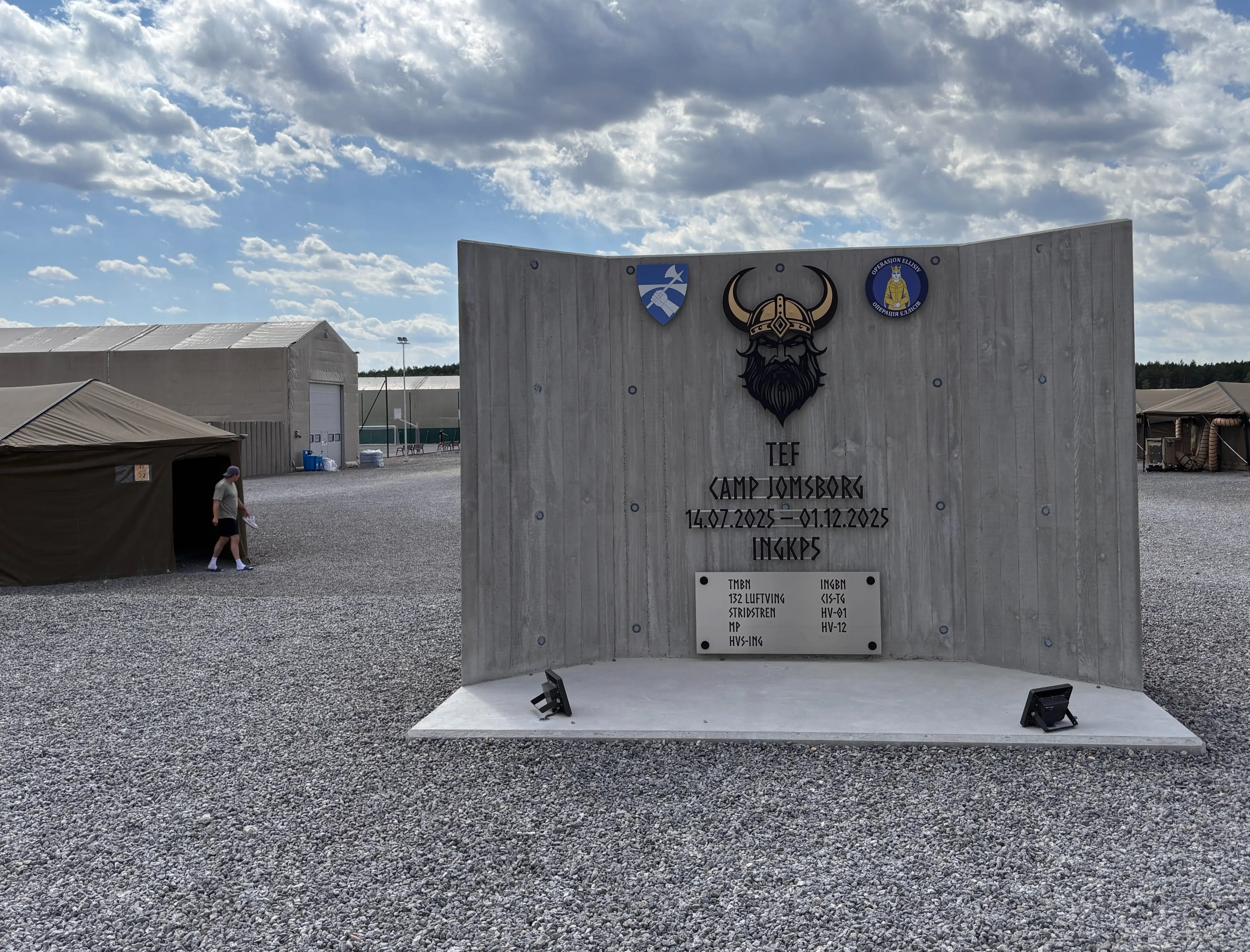 Concrete Camp Jomsborg sign with Viking emblem stands among tents on a gravel site under a cloudy sky.
